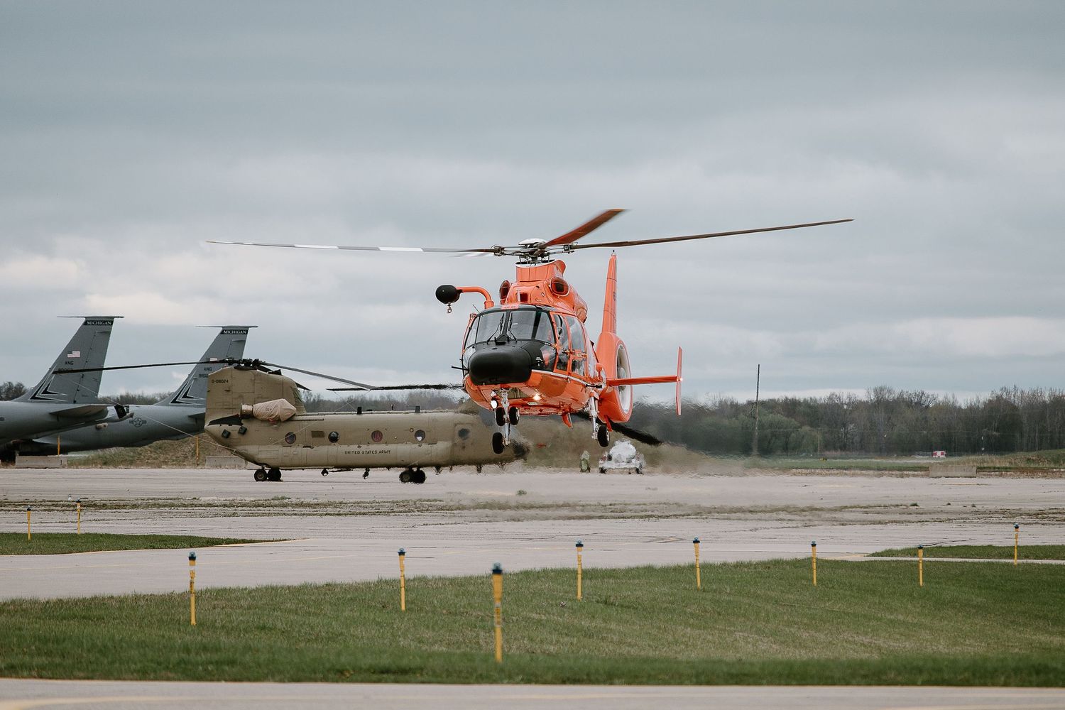 Military helicopters and aircraft lined up on airfield under cloudy sky.
