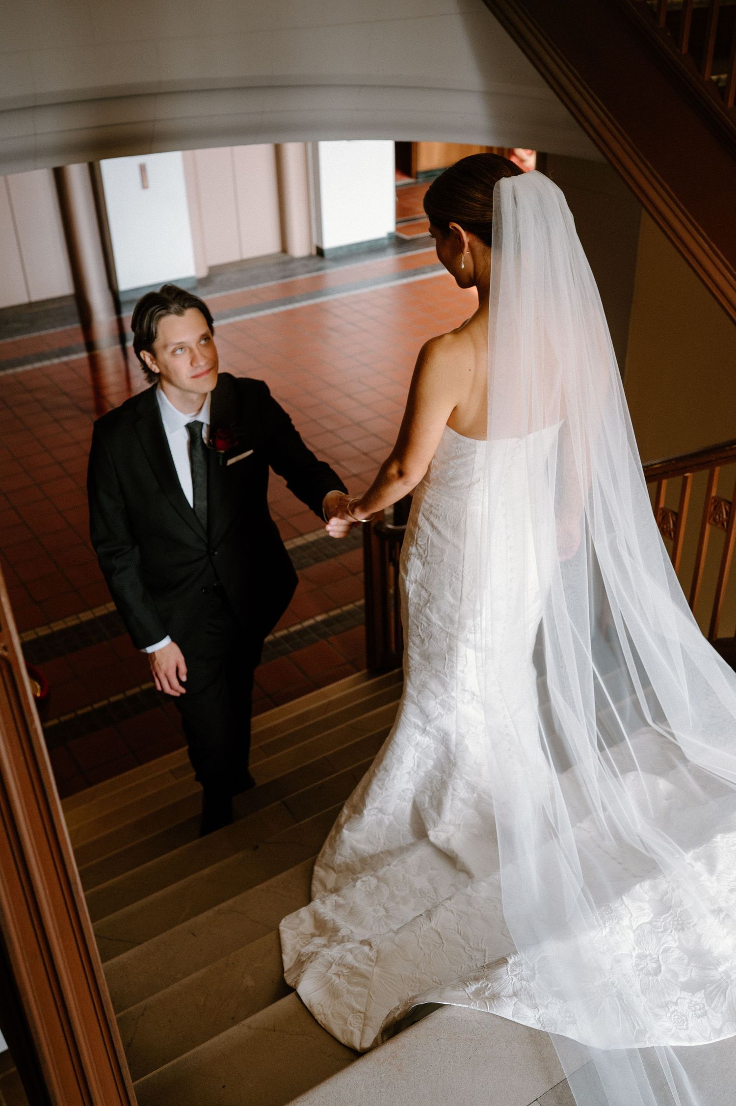 A bride in a flowing white gown with long veil meets her groom on an elegant wooden staircase.