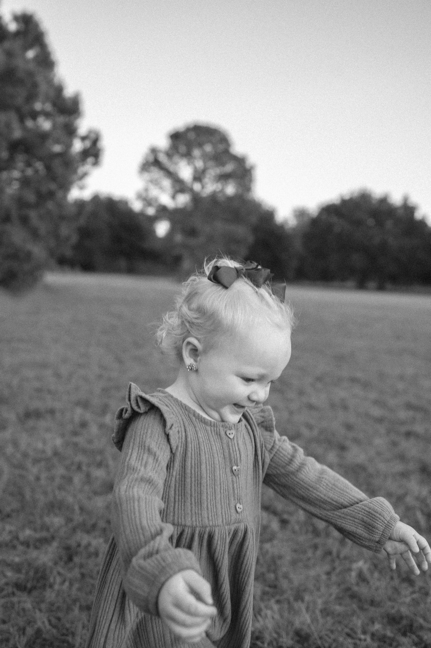 Black and white photo of a happy toddler girl in a ruffled dress with a bow, playing outdoors in a grassy park.