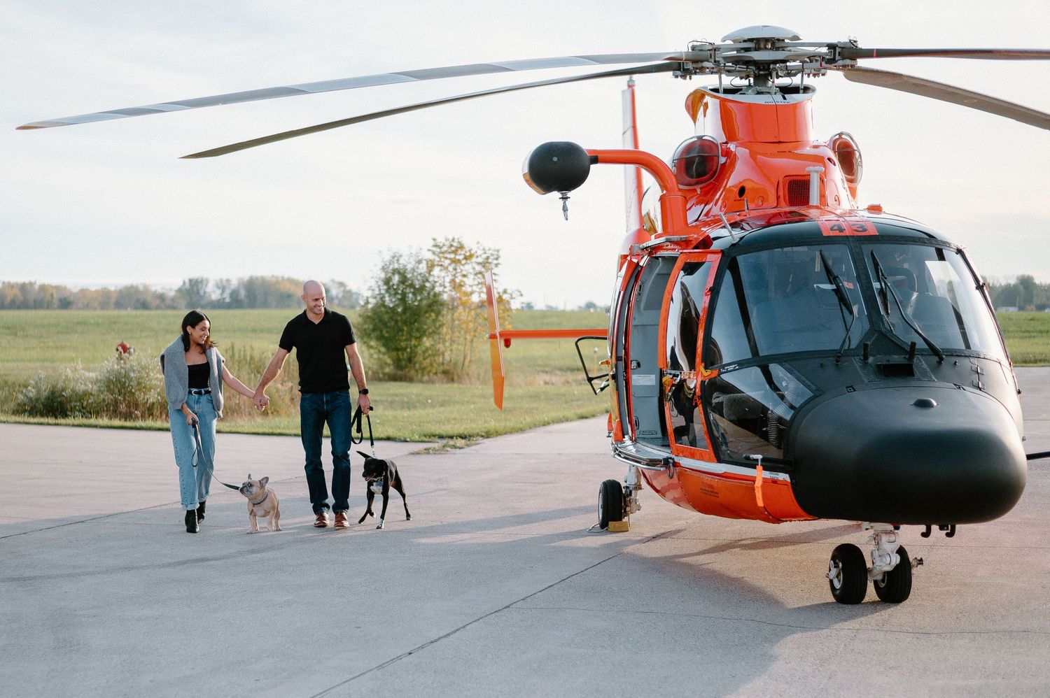 Large military helicopter sits on empty airfield with people walking nearby.