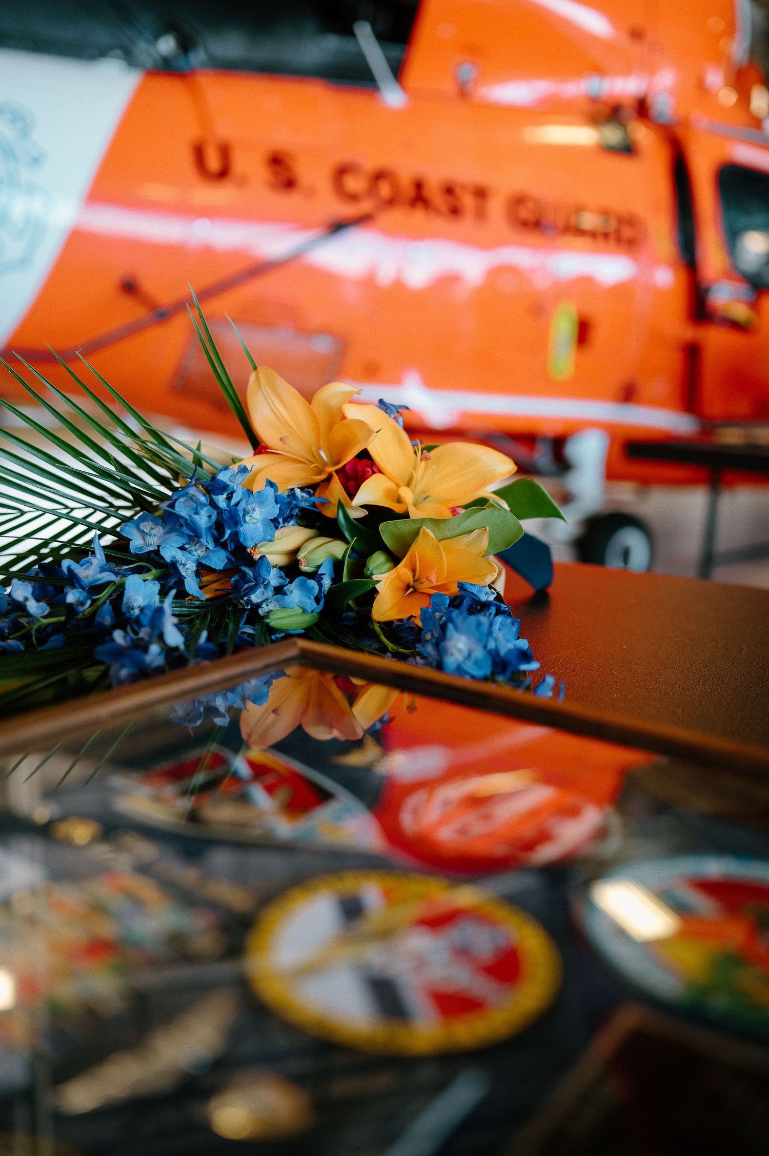 Blue and orange flowers rest on a reflective surface near a U.S. Coast Guard helicopter at a memorial ceremony.