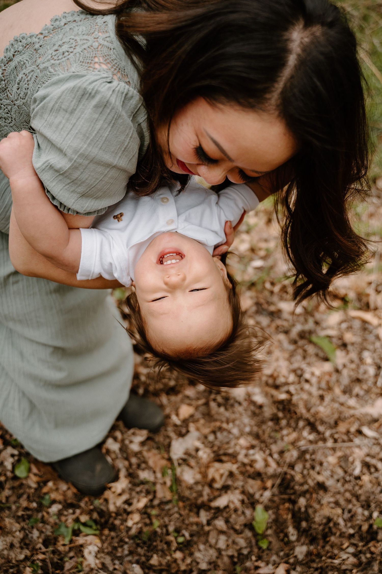 A joyful mother holds her laughing baby upside down outdoors among fallen leaves in a heartwarming candid moment.