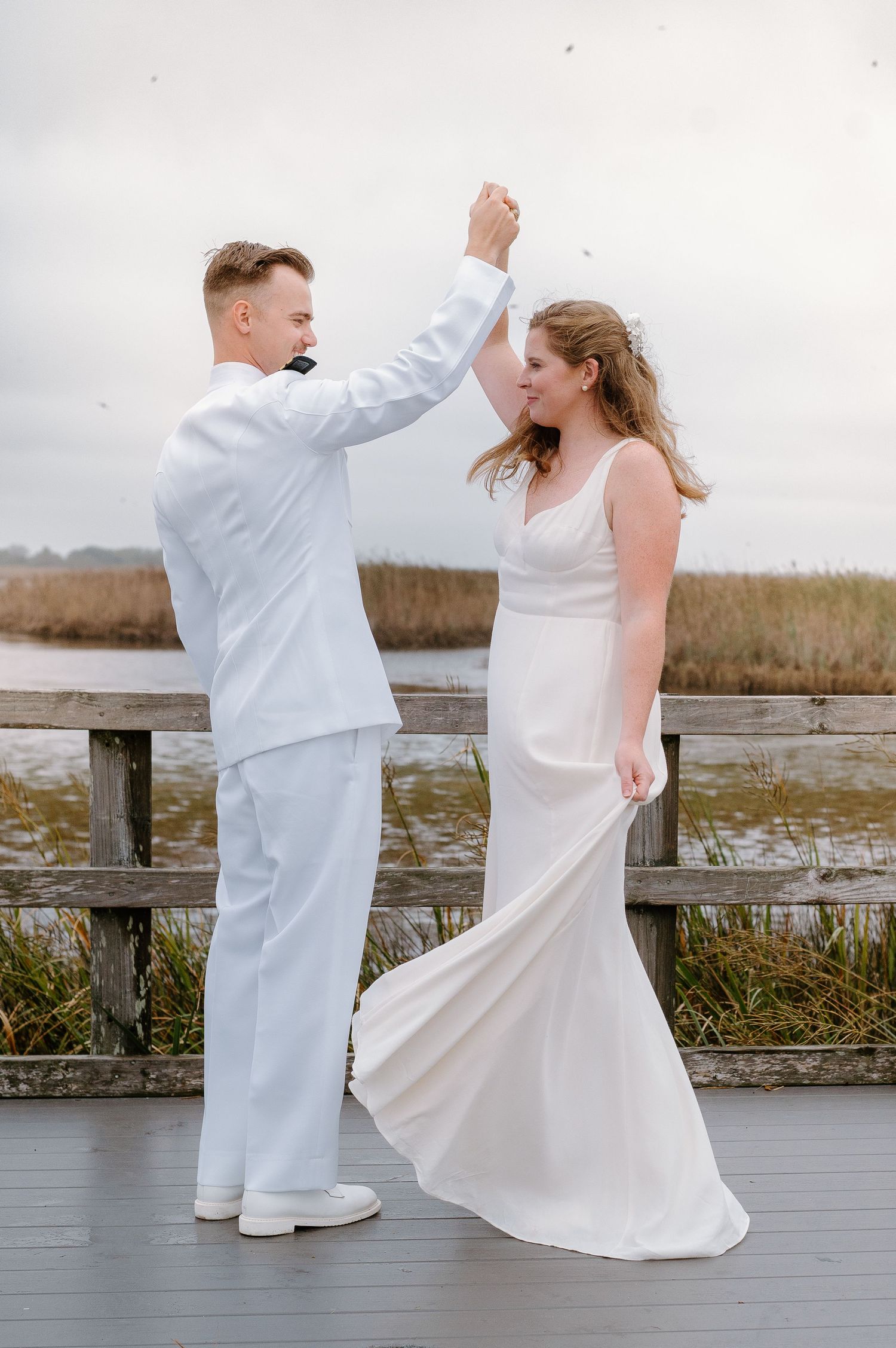 A couple in white attire dancing playfully on a waterfront boardwalk.