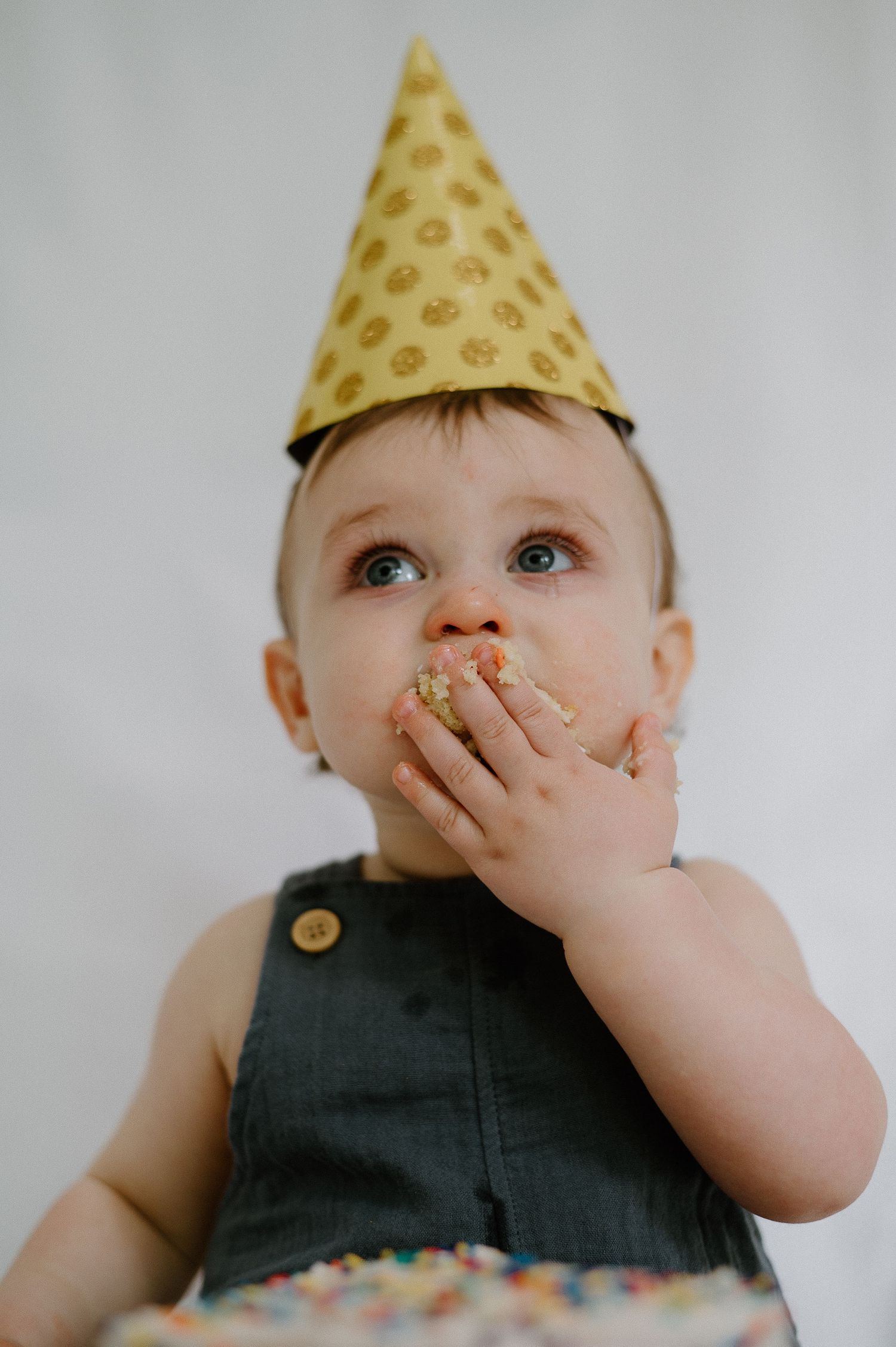 Baby wearing party hat and dark outfit covers mouth with hand while celebrating.