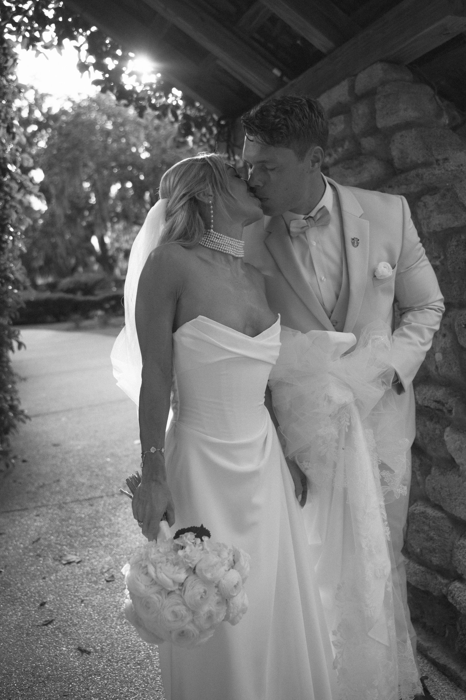 Black and white photo of a bride and groom sharing a kiss, bride holding a bouquet, outdoors near stone wall.