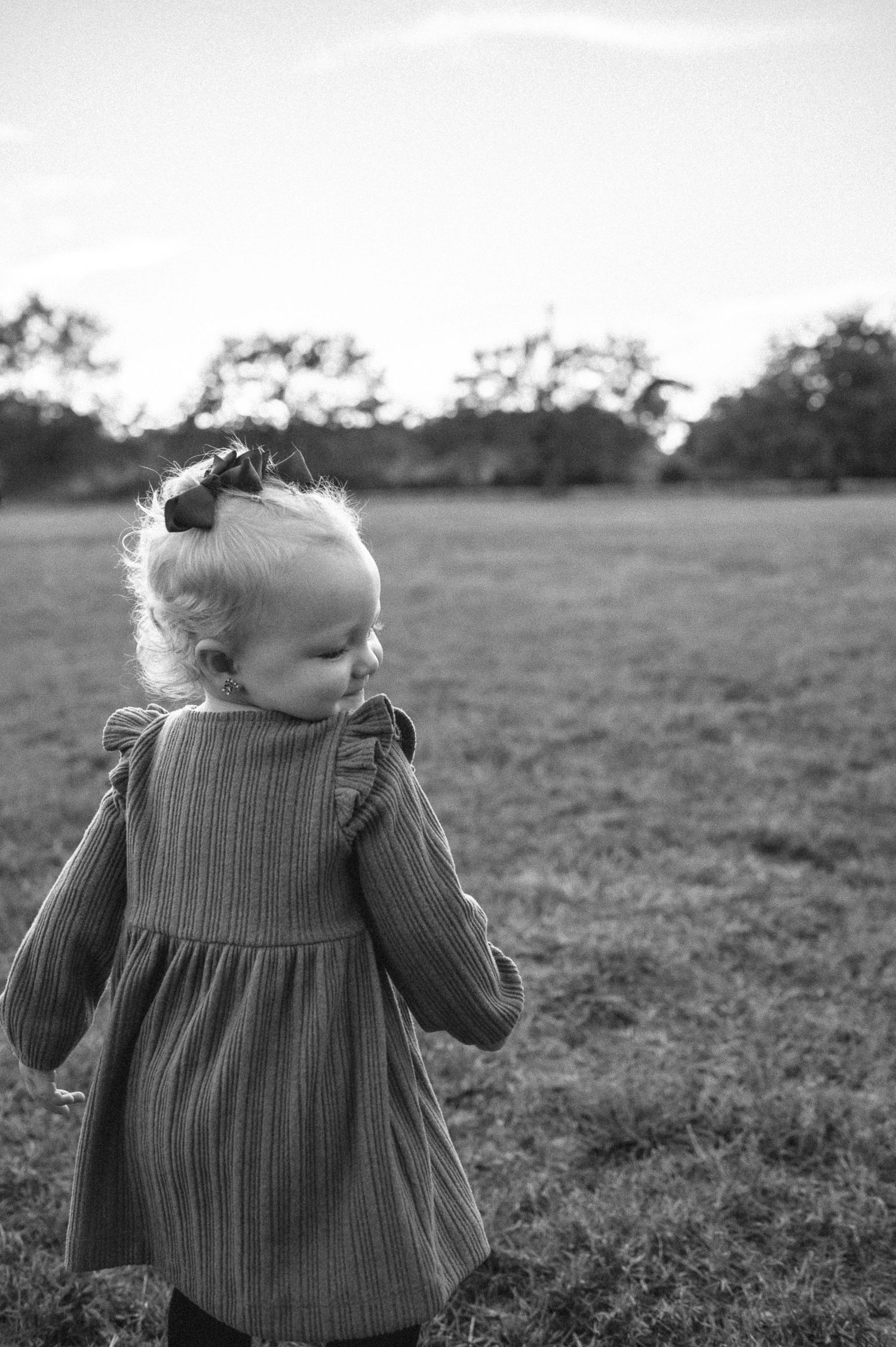 Black and white photo of a toddler girl in a ruffled dress standing in an open grassy field.