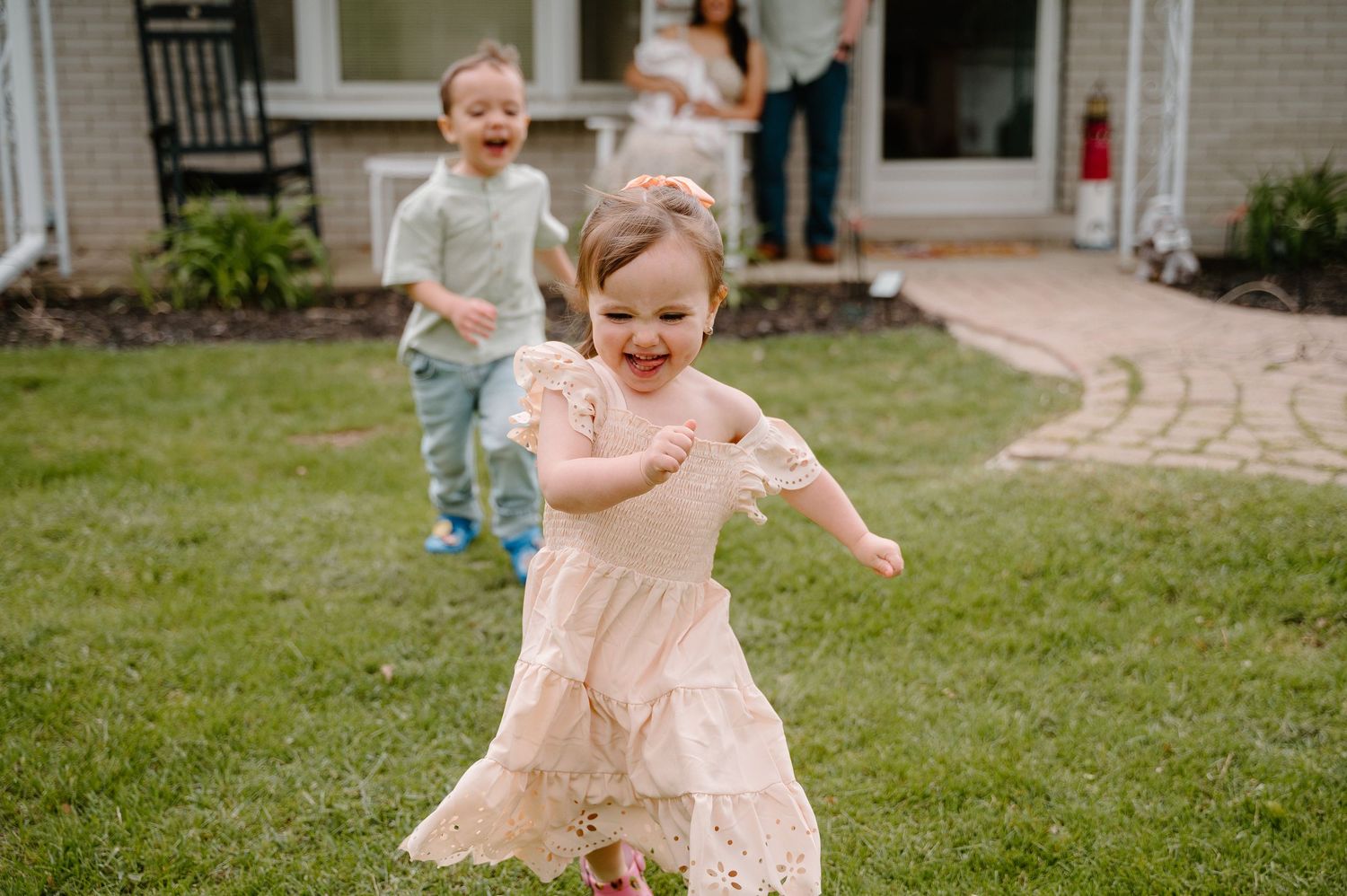 Two toddlers playing in a backyard, a girl in a pink dress running toward the camera while a boy laughs behind her.