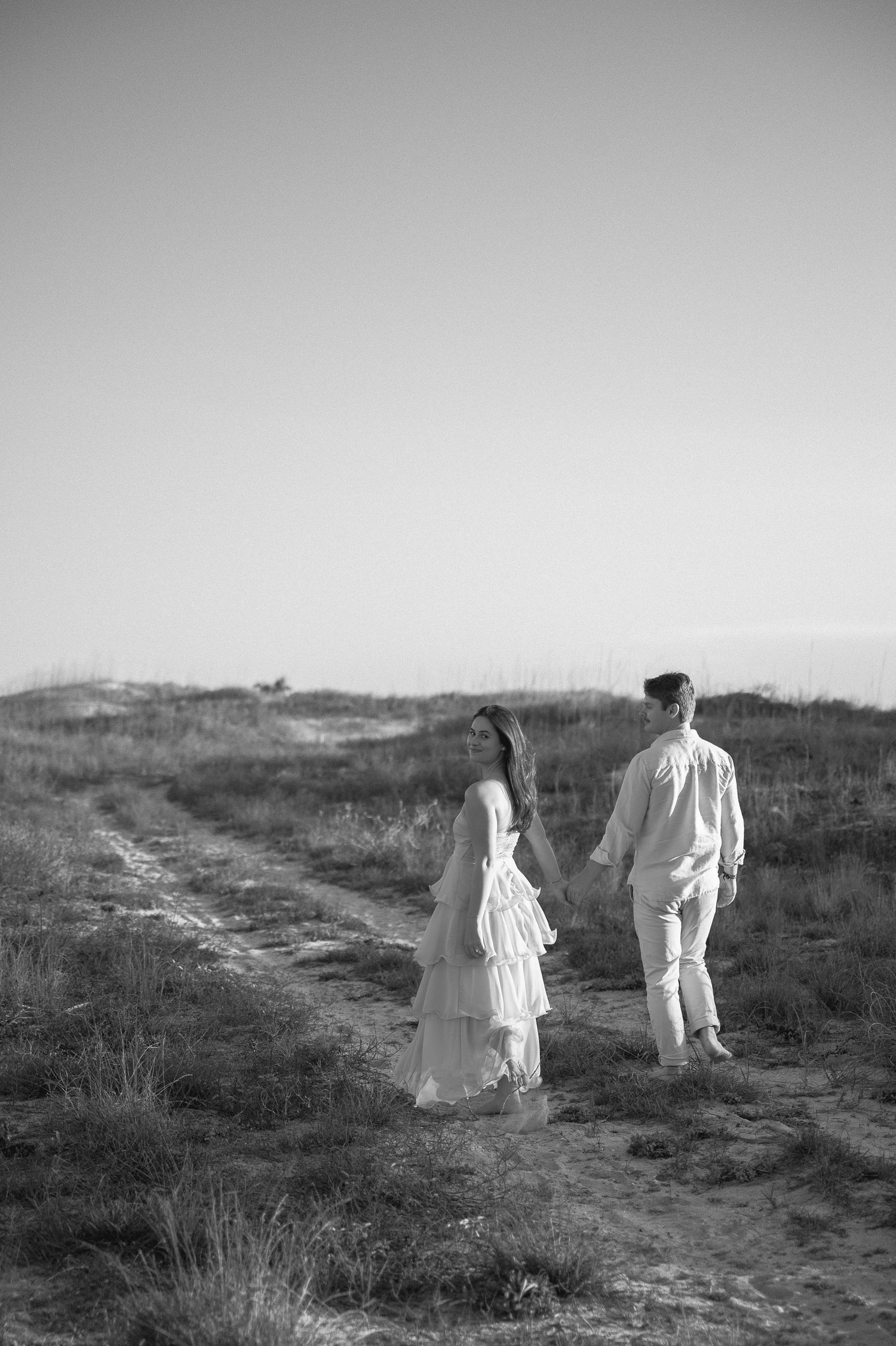 Two figures in white clothing walk along a winding dirt path through a misty field at dusk.