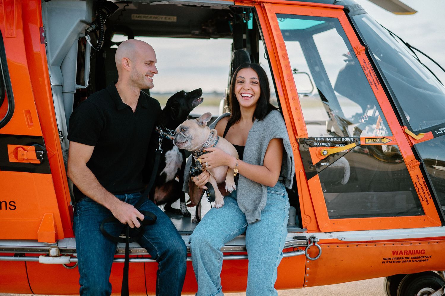 Family and pets gather for photos around bright orange helicopter on airfield.