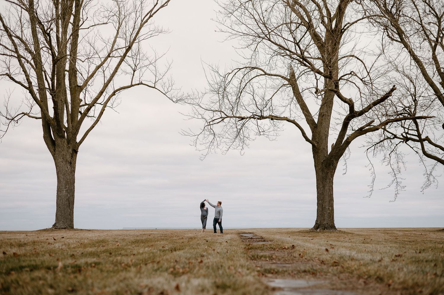 Silhouetted couple stands between bare winter trees in open field.