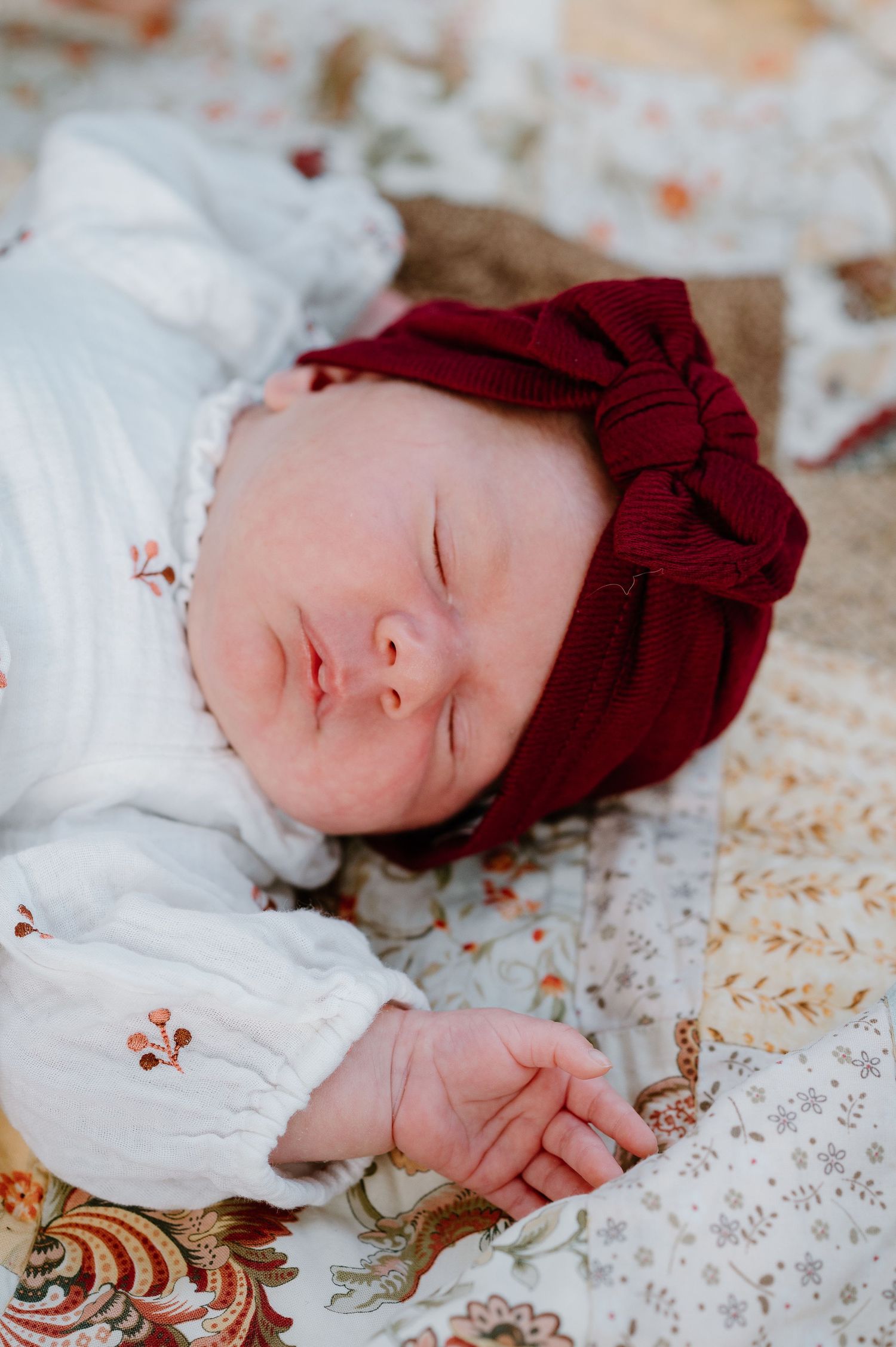 A sleeping baby in a burgundy bow hat and white dress rests on a floral print blanket.