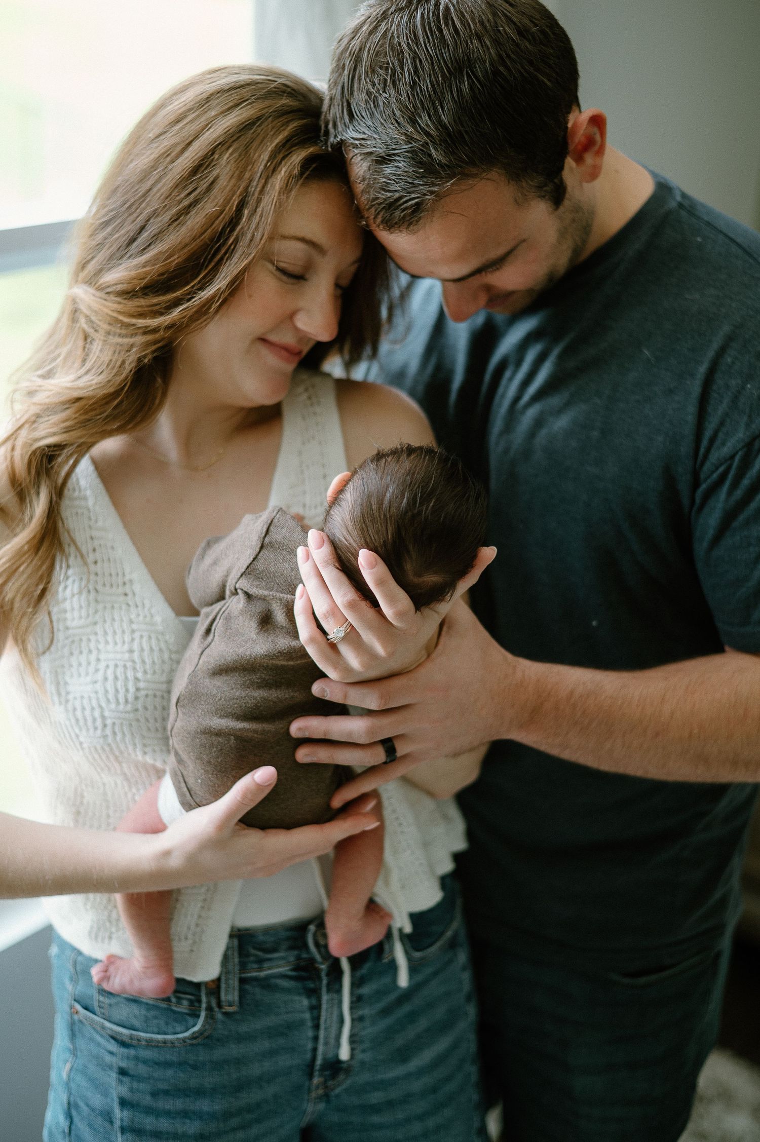 A couple gazes lovingly at their infant near a sunlit window.