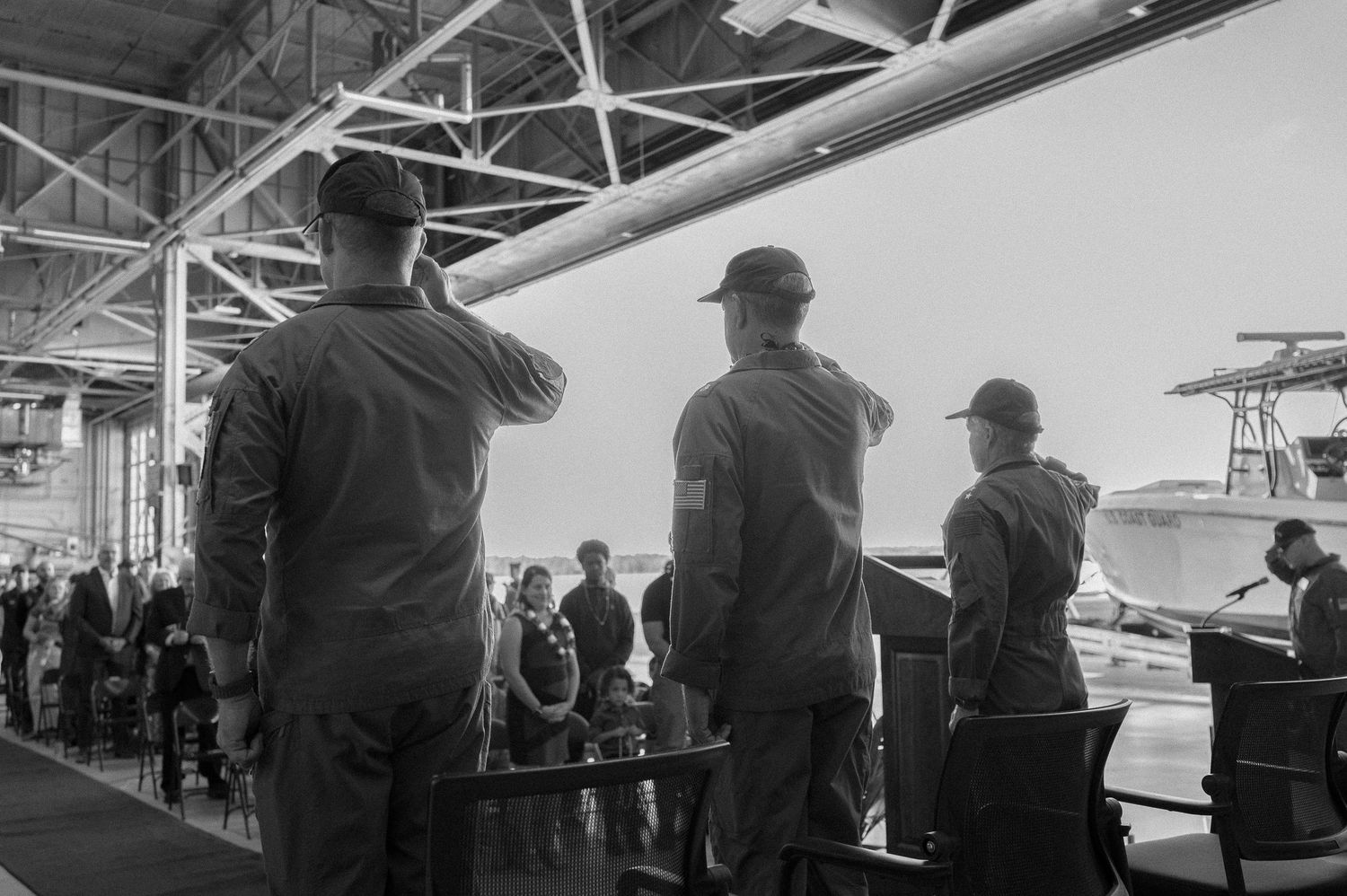 Military personnel saluting during a formal ceremony inside a hangar with boats visible in the background.