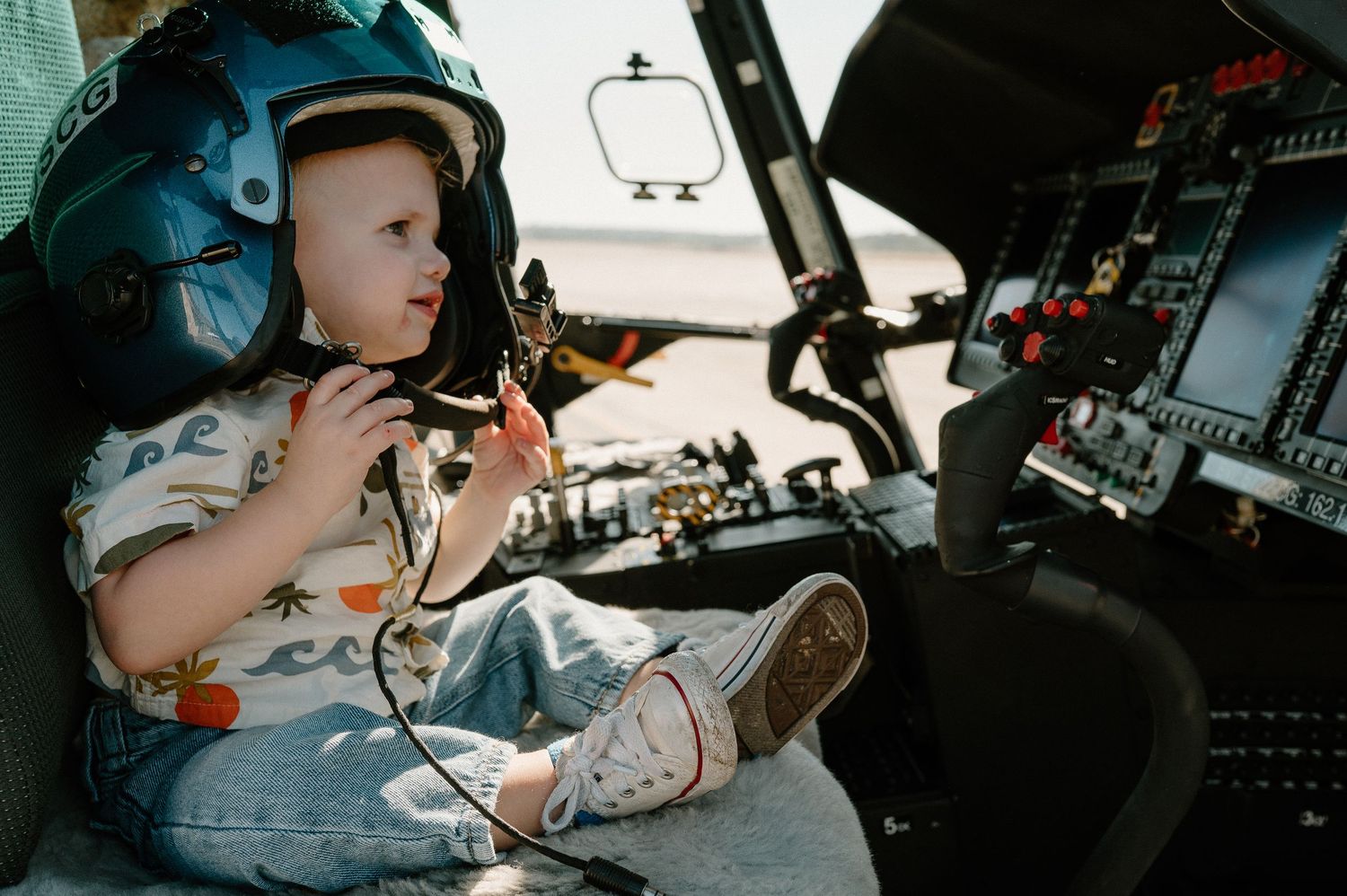 Young child wearing a pilot helmet sits in a helicopter cockpit, exploring the controls and instruments.