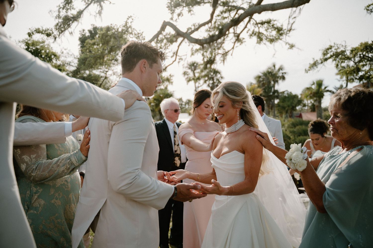 Outdoor wedding ceremony with bride and groom exchanging vows surrounded by guests under a tree.