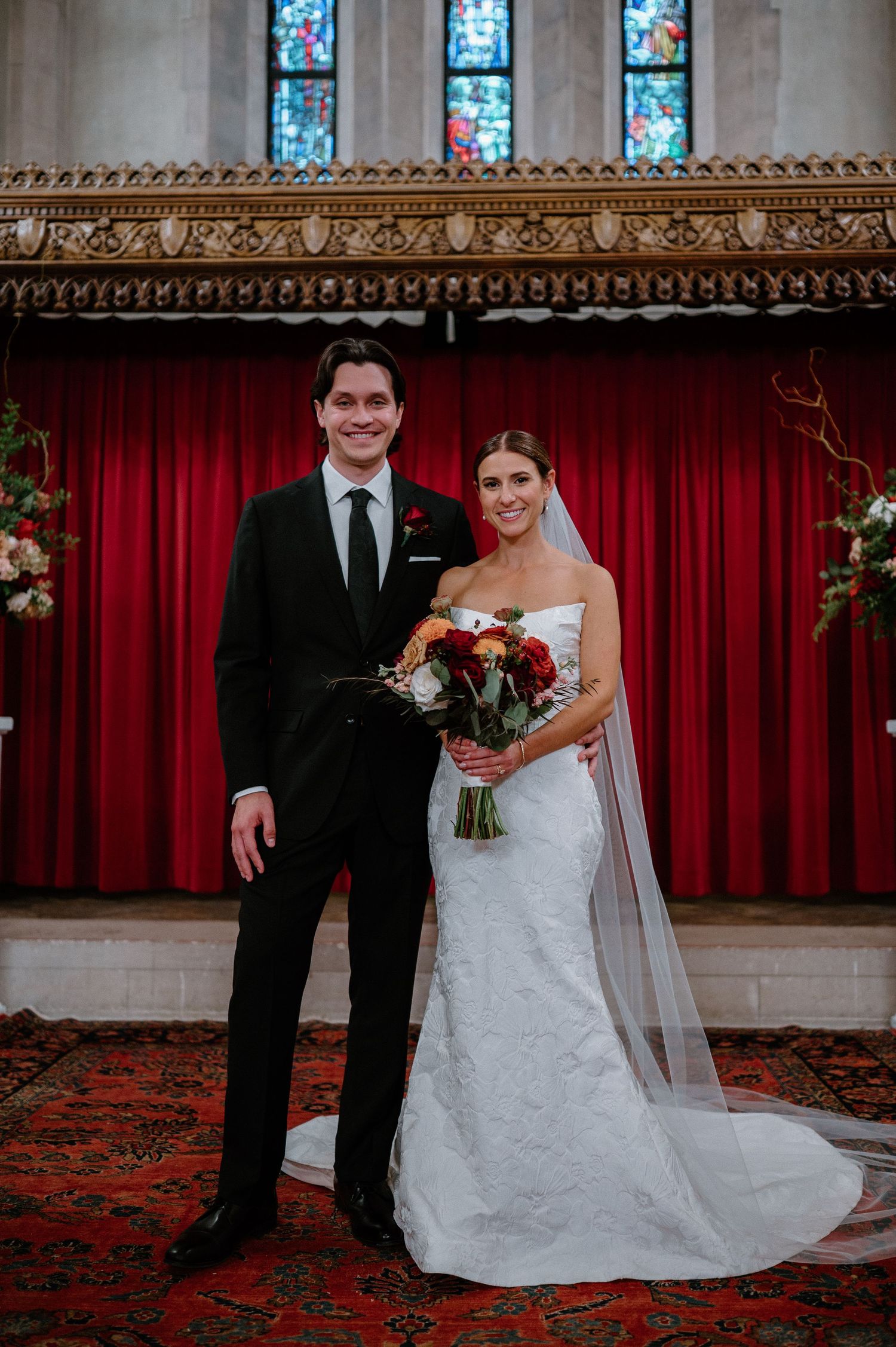 Bride and groom pose together in church with stained glass windows and red carpet in a series of wedding portraits.