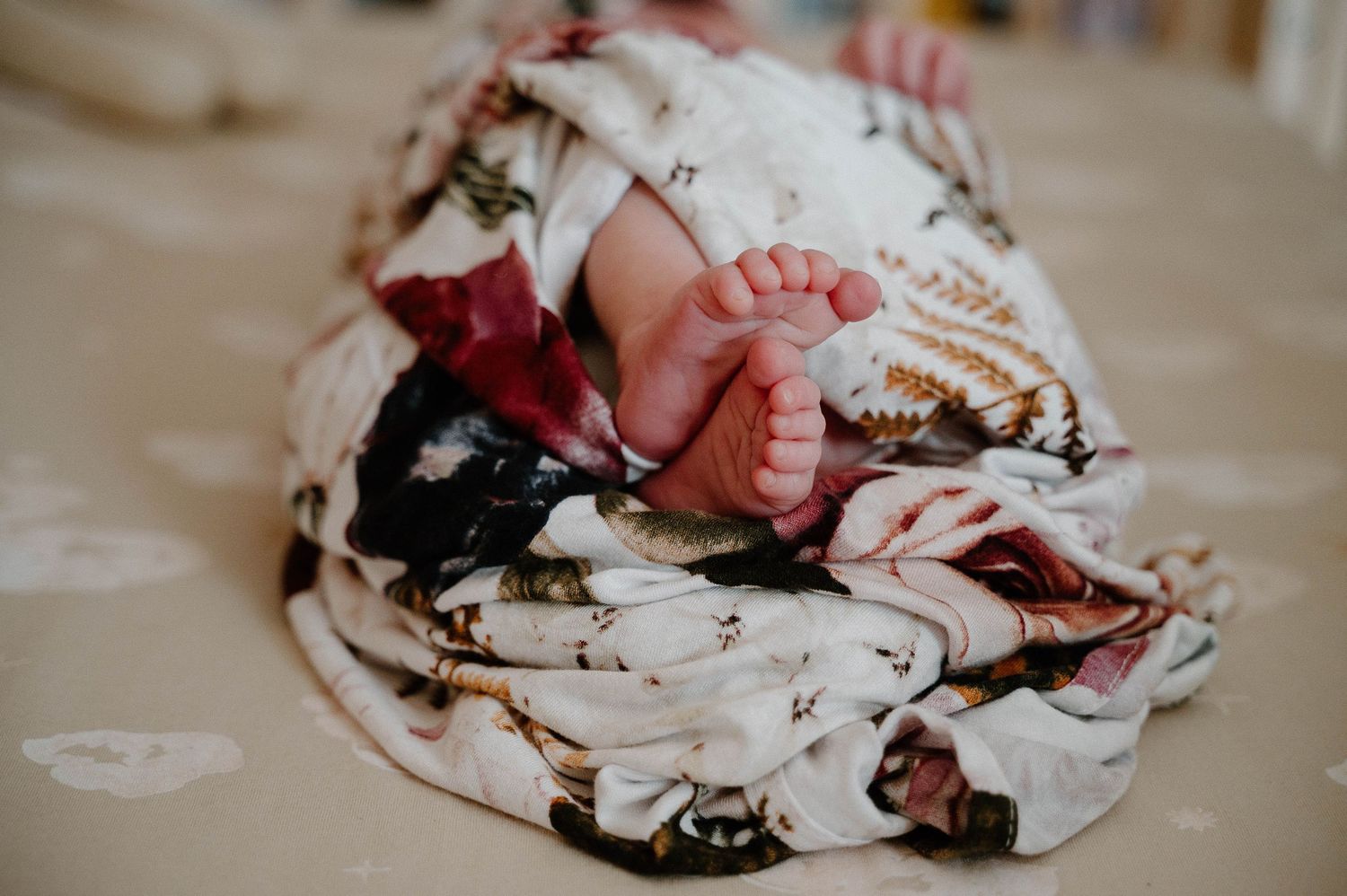Newborn baby swaddled in a floral muslin blanket with tiny feet peeking out, resting on a soft surface.