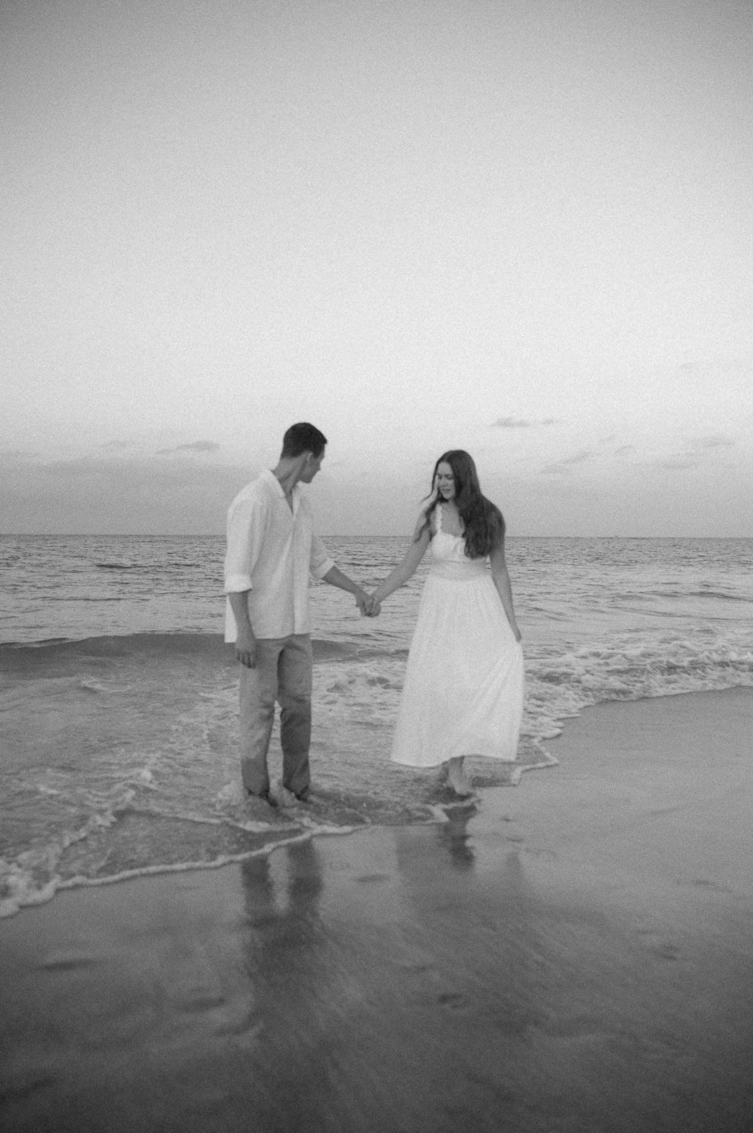 A couple holds hands on a beach at sunset in a romantic black and white photo, woman in white dress.