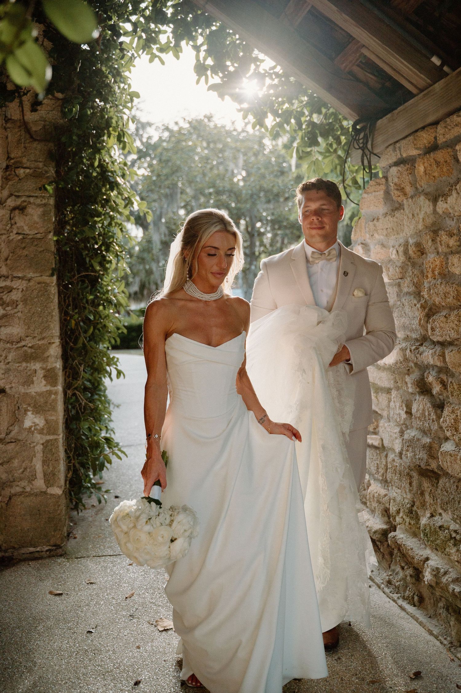 Bride in strapless white gown holding bouquet walks with groom in tan suit through stone archway with lush greenery.