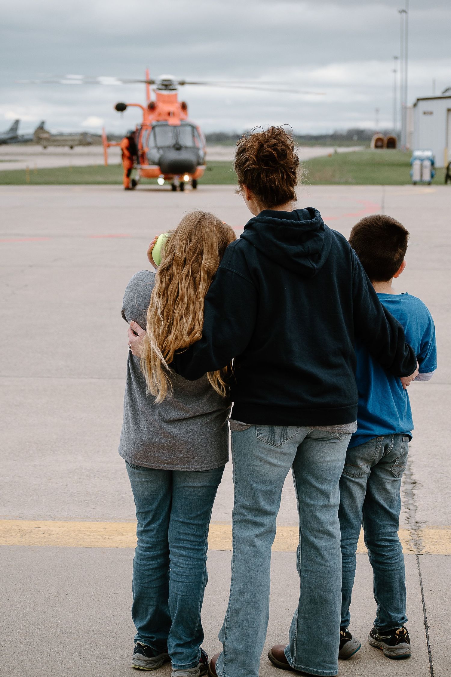 Three people watch an orange Coast Guard helicopter on the tarmac.
