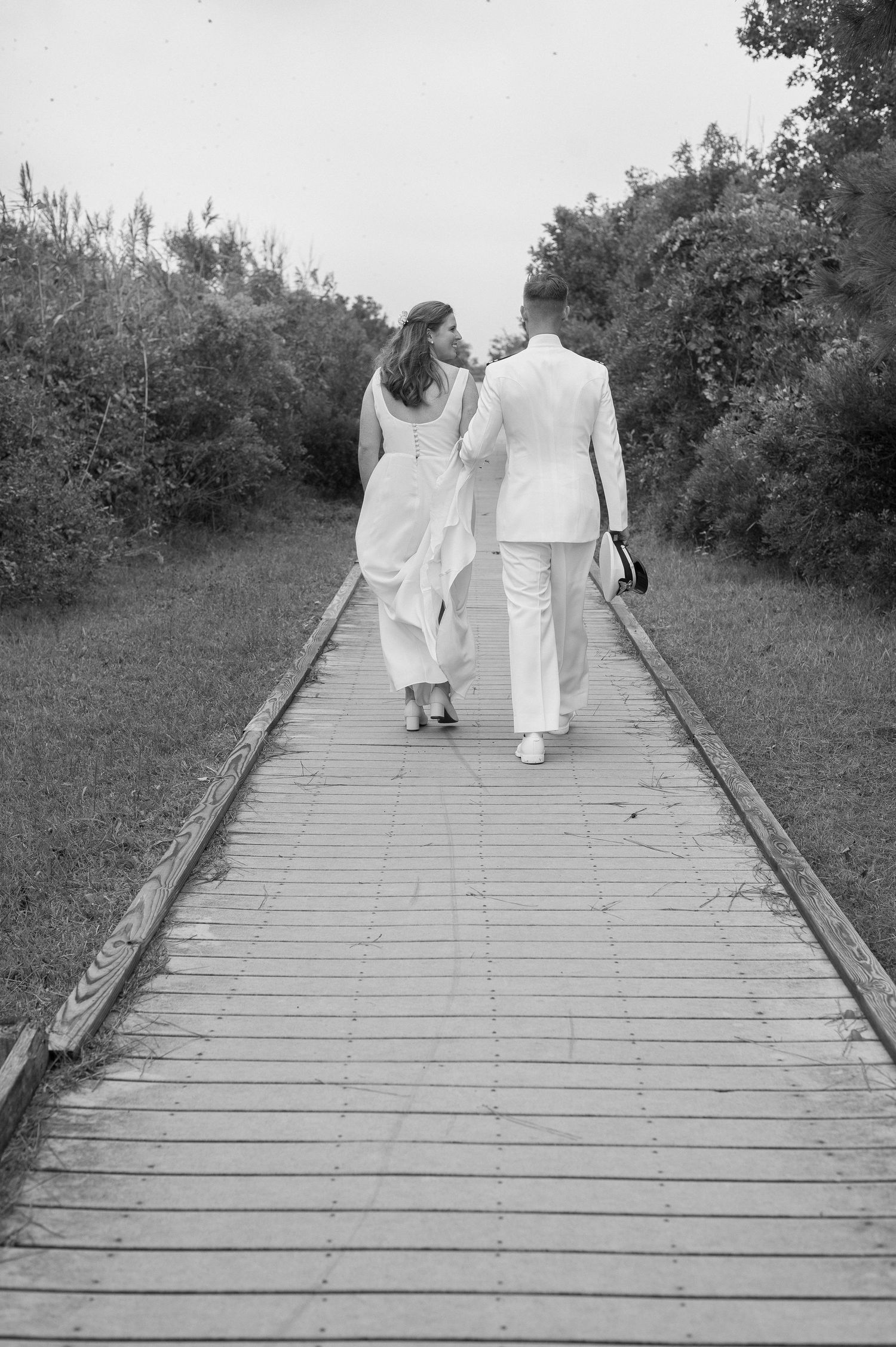 Two figures in white clothing walk along a wooden boardwalk surrounded by greenery.