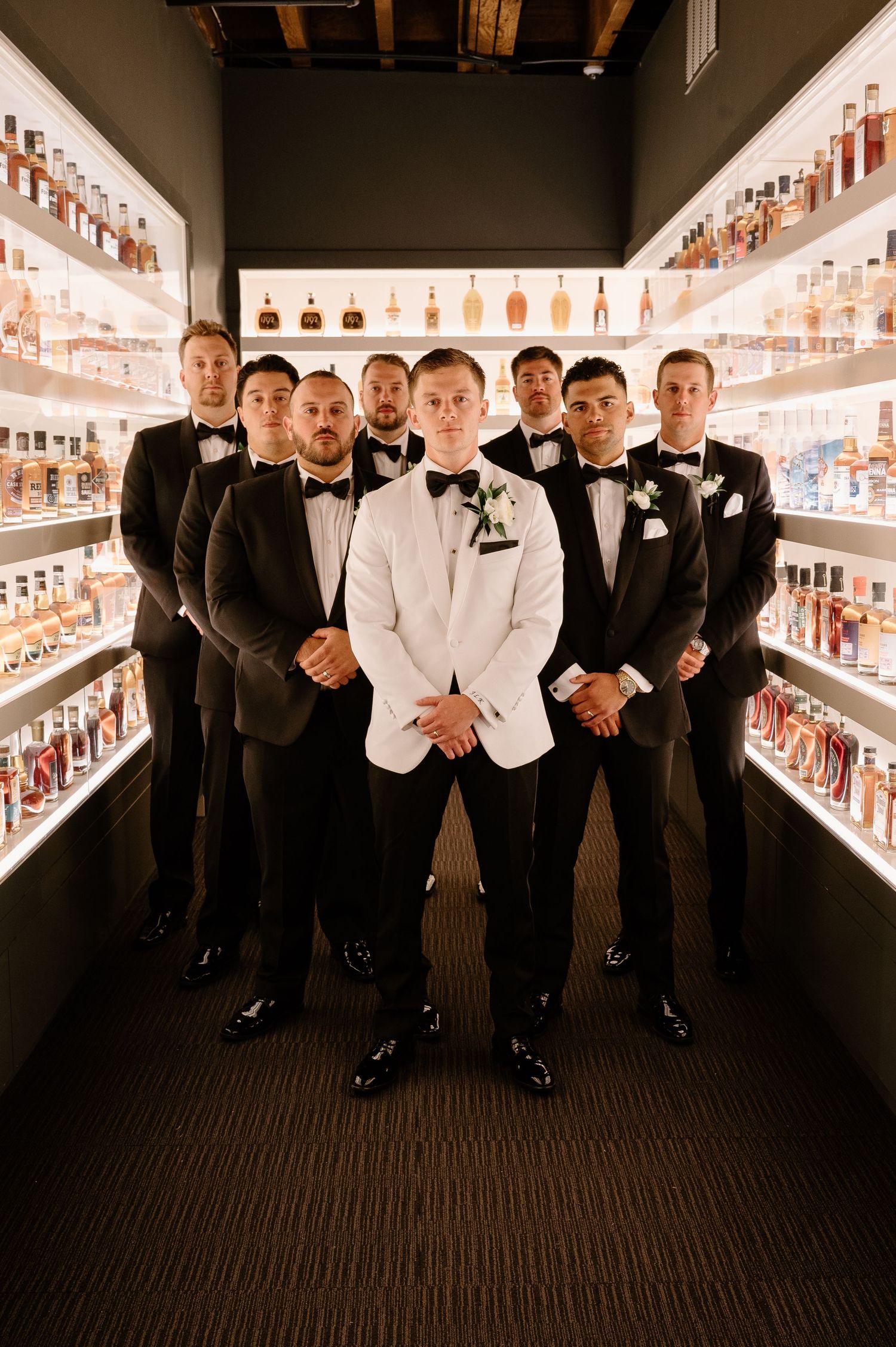 Groomsmen in formal attire posing in front of a wall of liquor bottles in a venue.