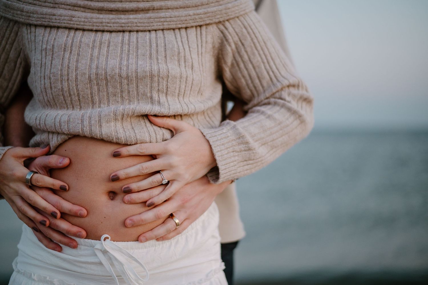 Person in beige sweater holding their stomach against oceanic backdrop.