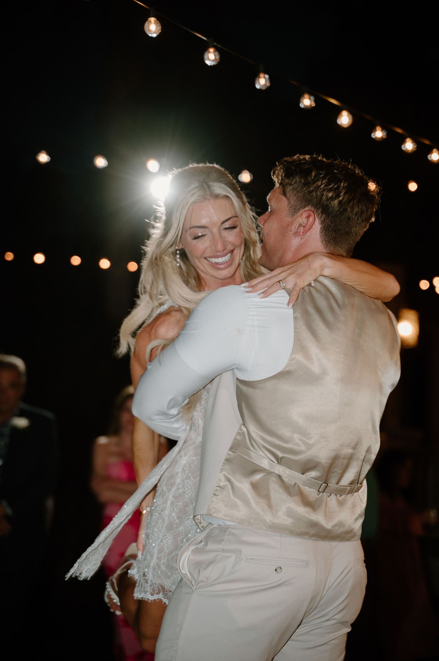 A joyful bride in white is lifted by her groom in a gold vest during an outdoor wedding reception under string lights.