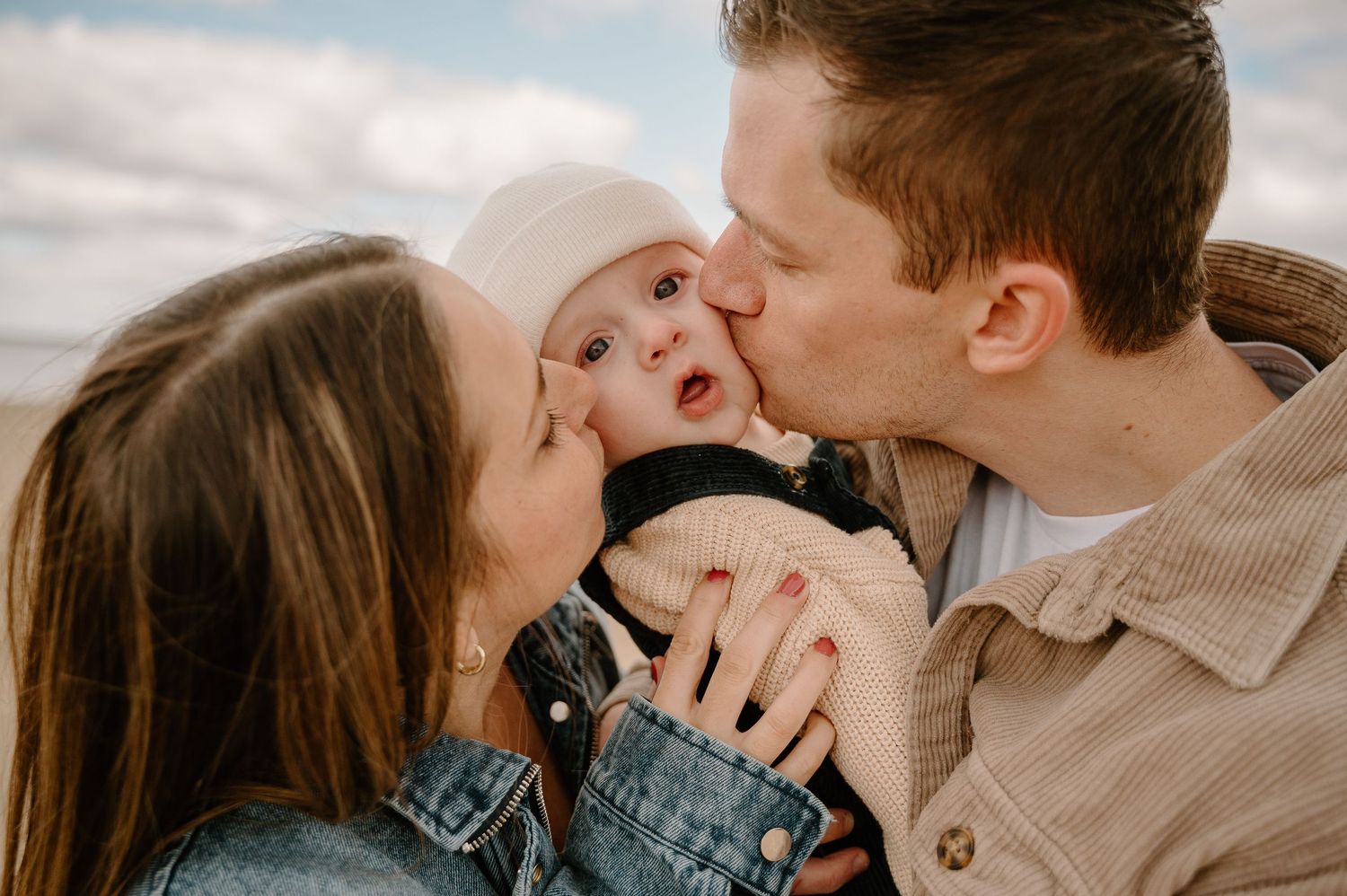Two parents kissing their adorable baby on the cheeks outdoors on a cloudy day.