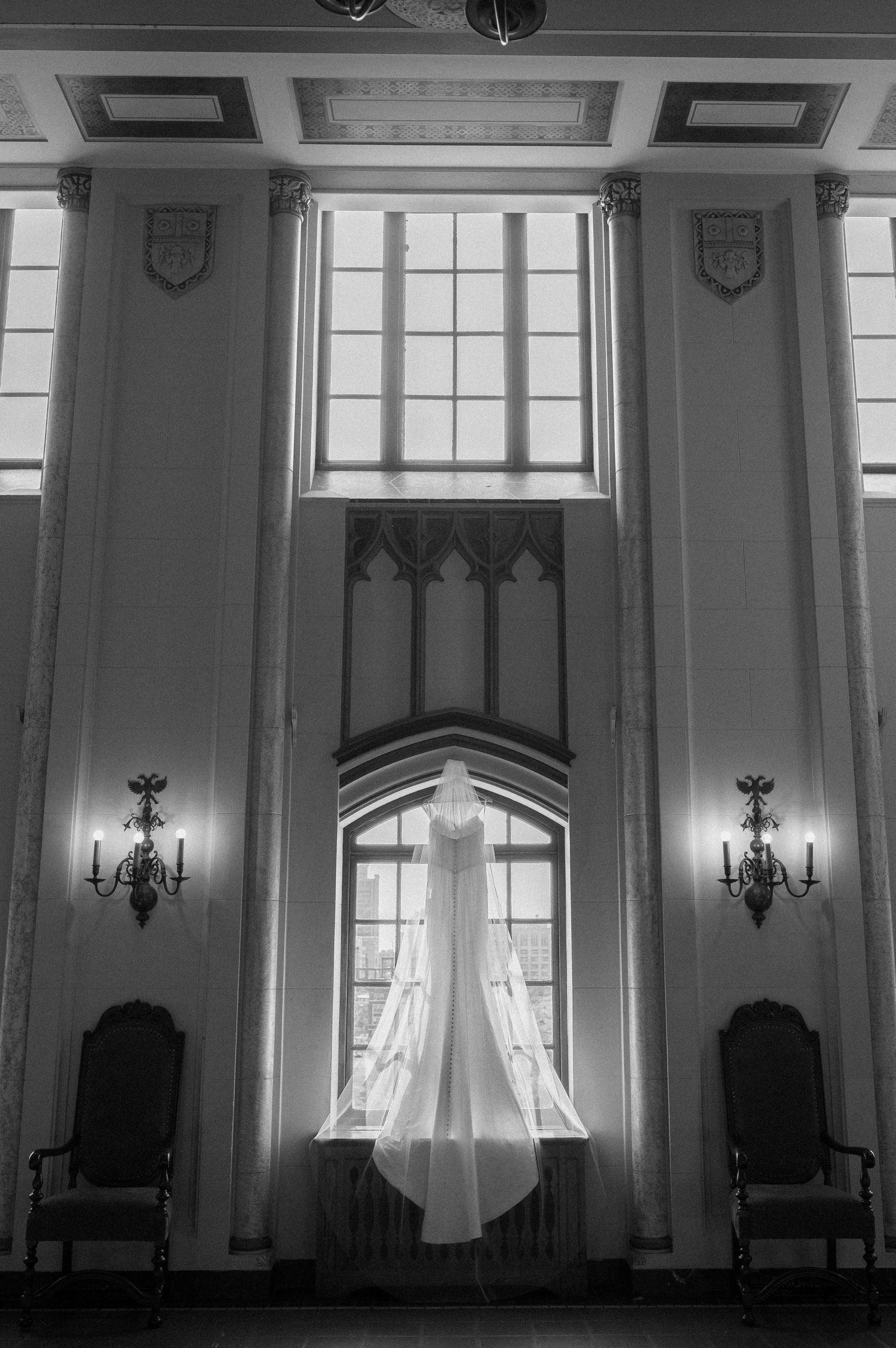 A white wedding dress hangs elegantly in a grand room with ornate wall sconces and high ceilings.