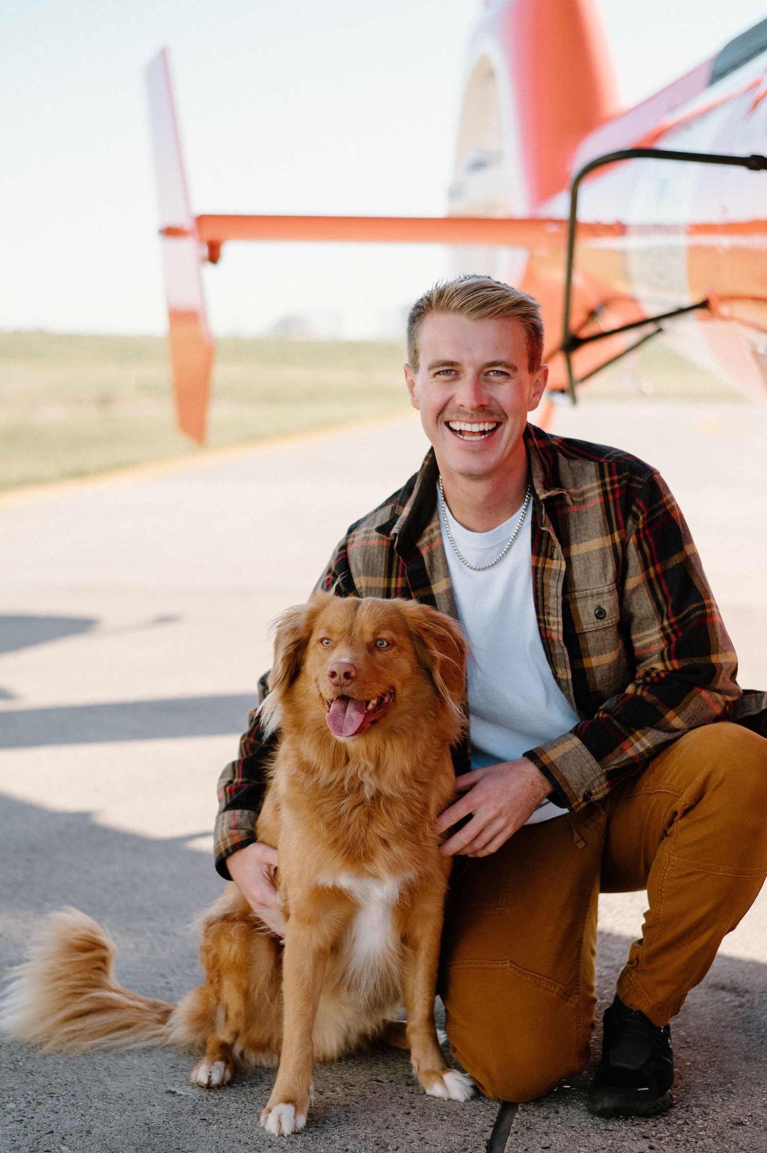 Golden retriever and friend sit together on airfield next to bright orange helicopter.