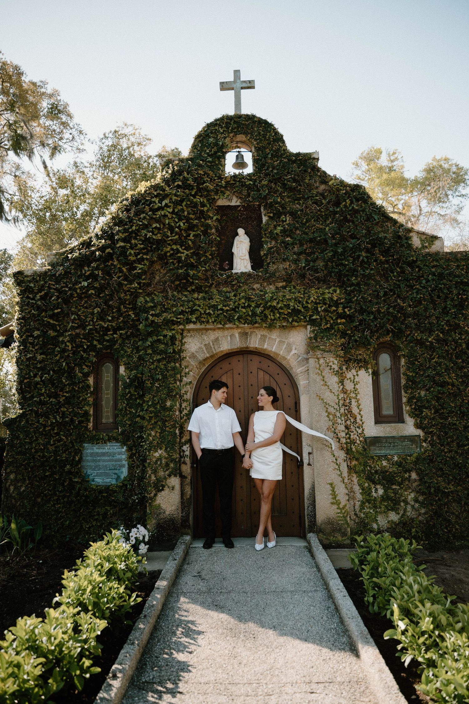 Couple holding hands outside an ivy-covered chapel with a cross, bell, and wooden arched door on a sunny day. our lady of la Leche shrine
