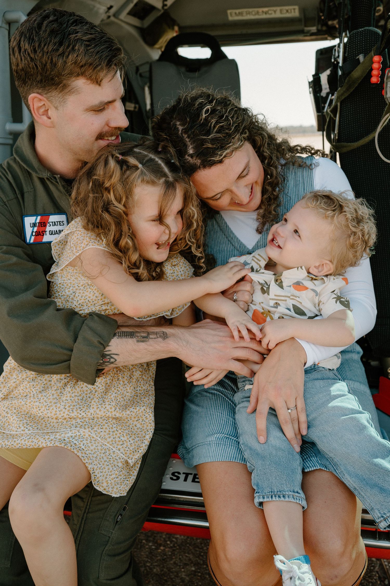 A happy family of four laughs together sitting at the back of a vehicle, parents hugging two young curly-haired children.