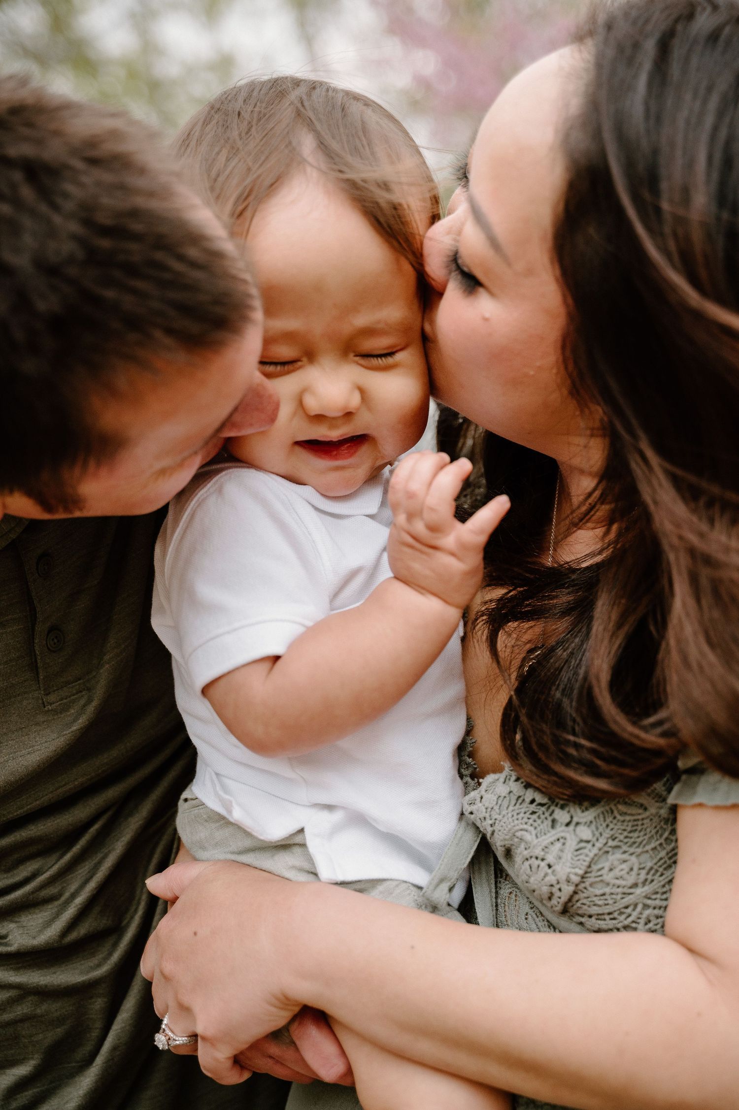 Parents kissing their laughing baby on both cheeks outdoors during a spring family photo session.