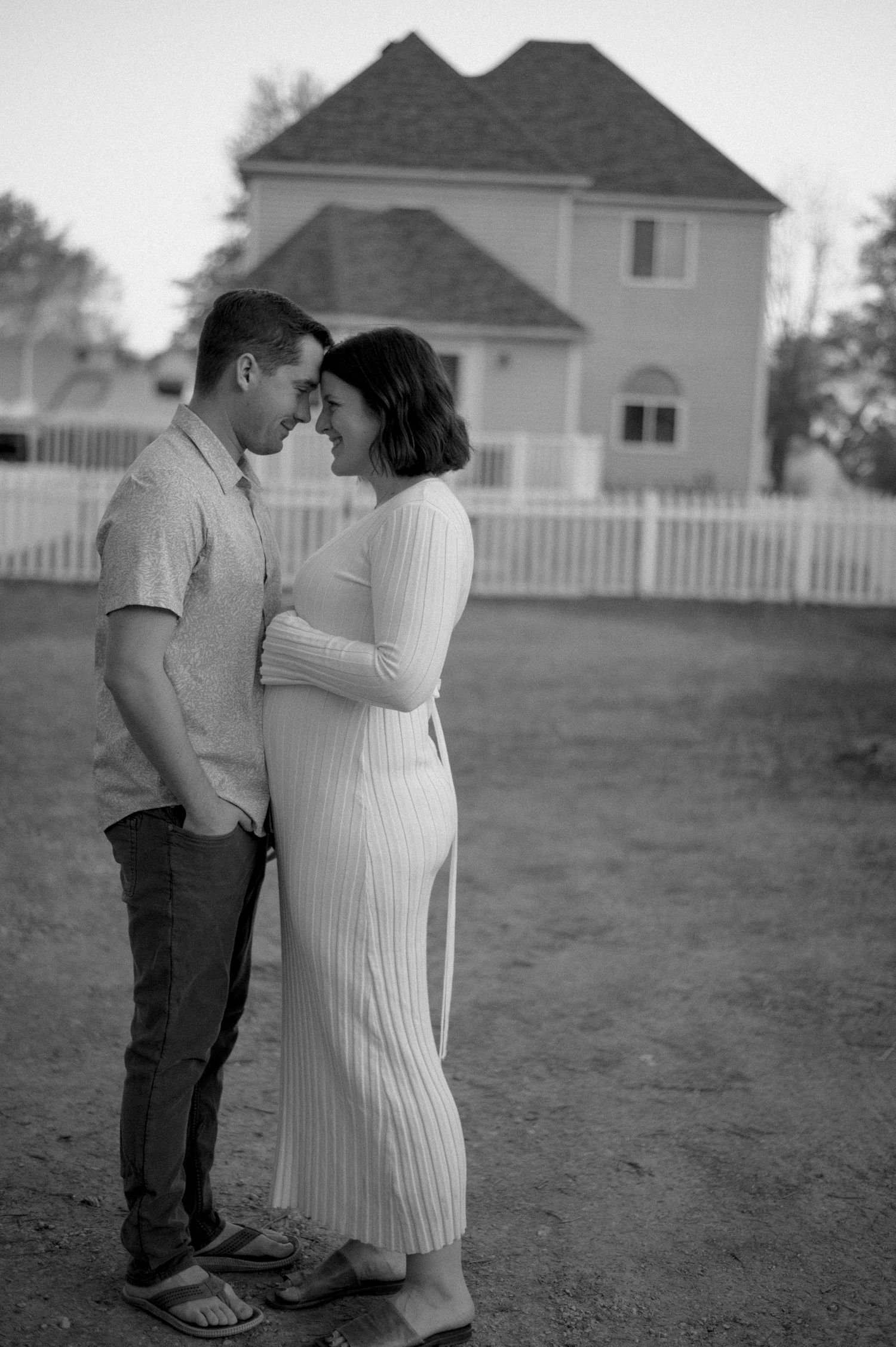 Black and white photo of a couple embracing outdoors near a white fence and house, the woman wearing a white ribbed dress.
