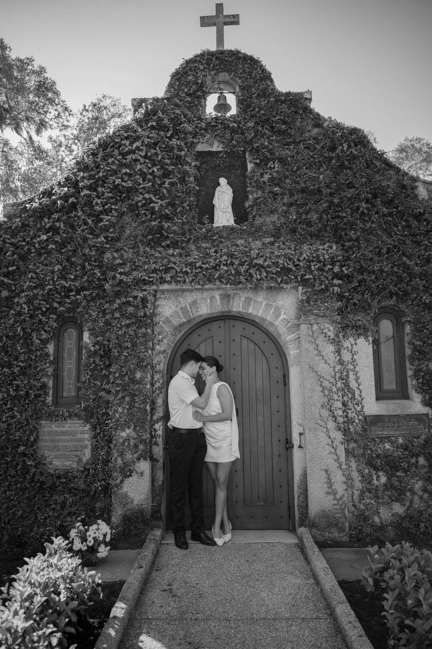 Black and white photo of a couple embracing before an ivy-covered chapel with a cross, bell, and statue above the arched door.
