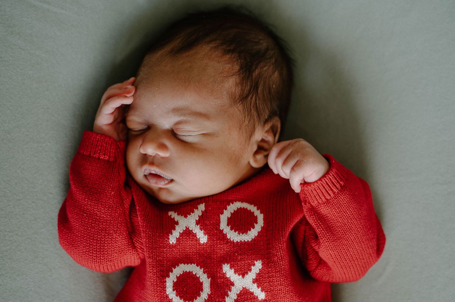 Sleeping newborn baby wearing a red knit XO sweater, lying on a soft gray background.
