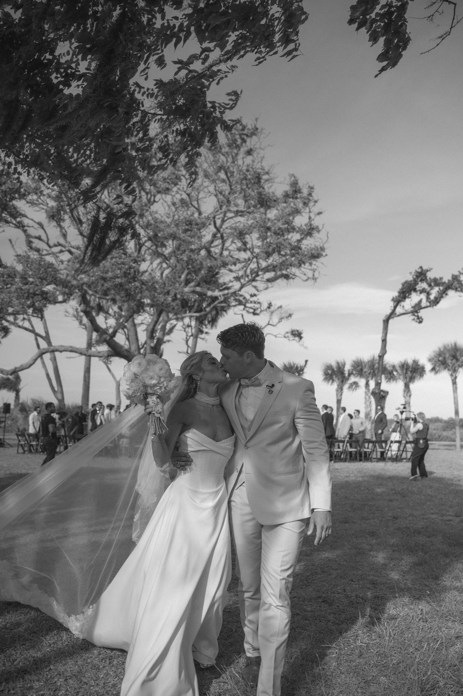 Black and white photo of a bride and groom sharing a kiss outdoors after their wedding ceremony, veil blowing in the wind.