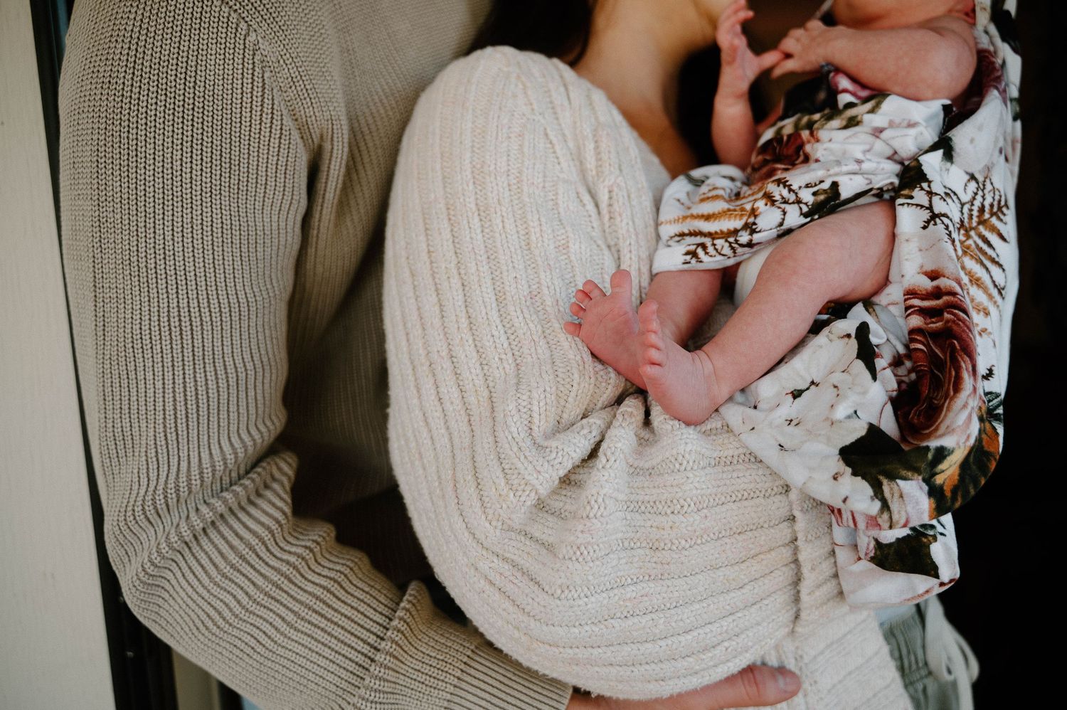 A parent in a cream knit sweater tenderly holds a newborn baby wrapped in a floral blanket.