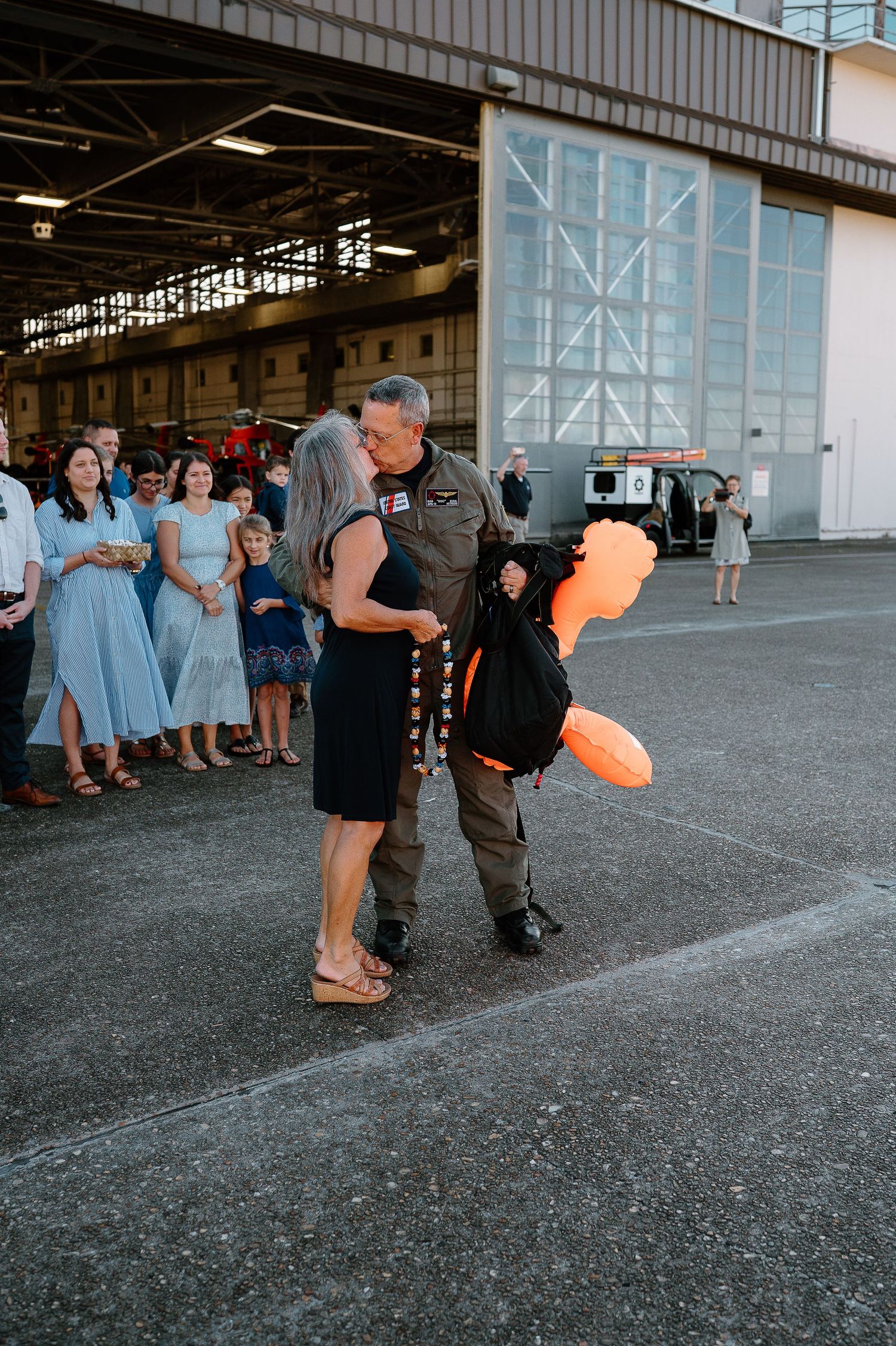 Military pilot in flight suit kisses woman while holding orange balloon animal outside hangar with crowd watching.