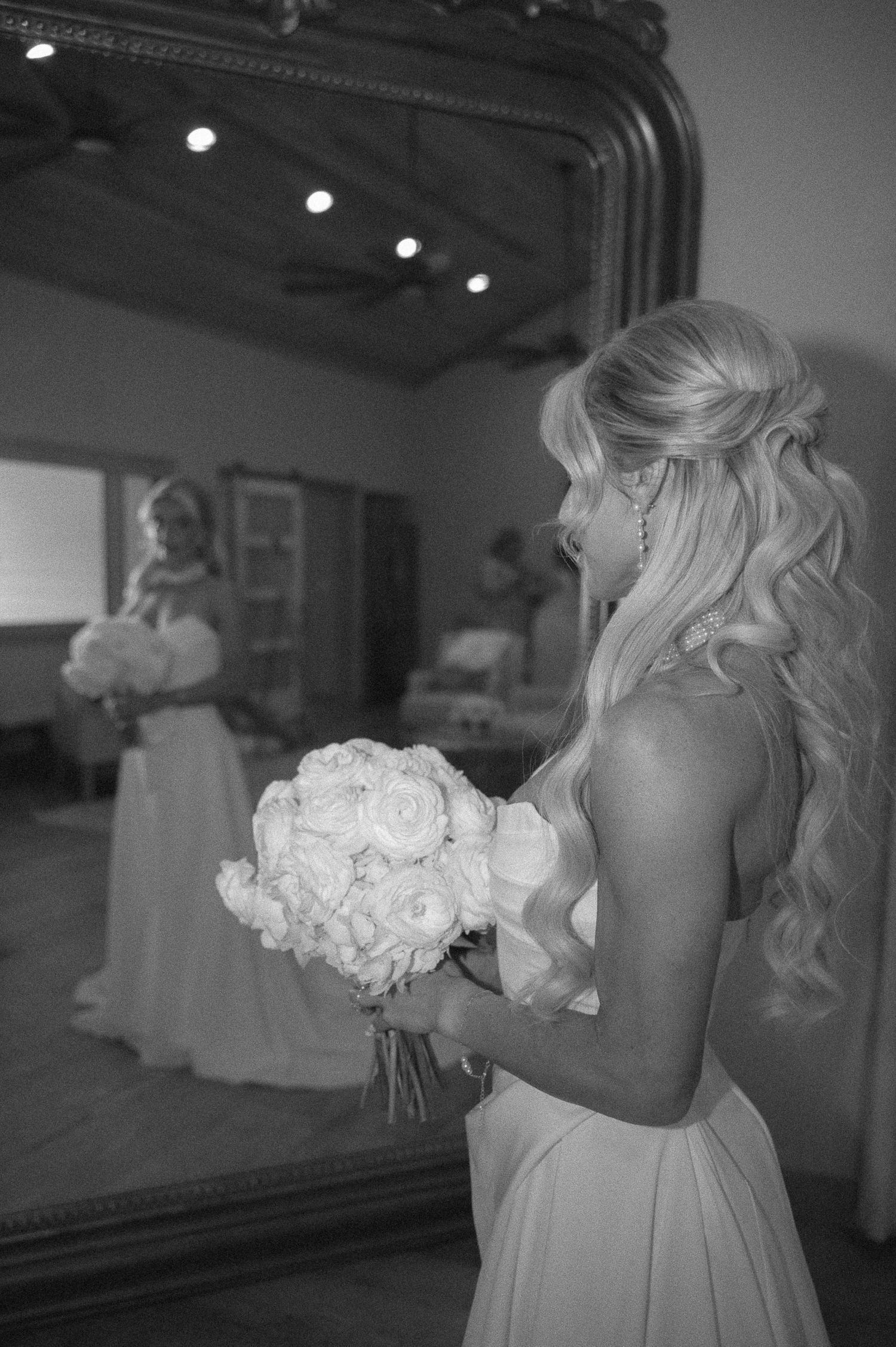 Bride in white gown holding bouquet of white roses, reflected in ornate mirror, black and white wedding photo.