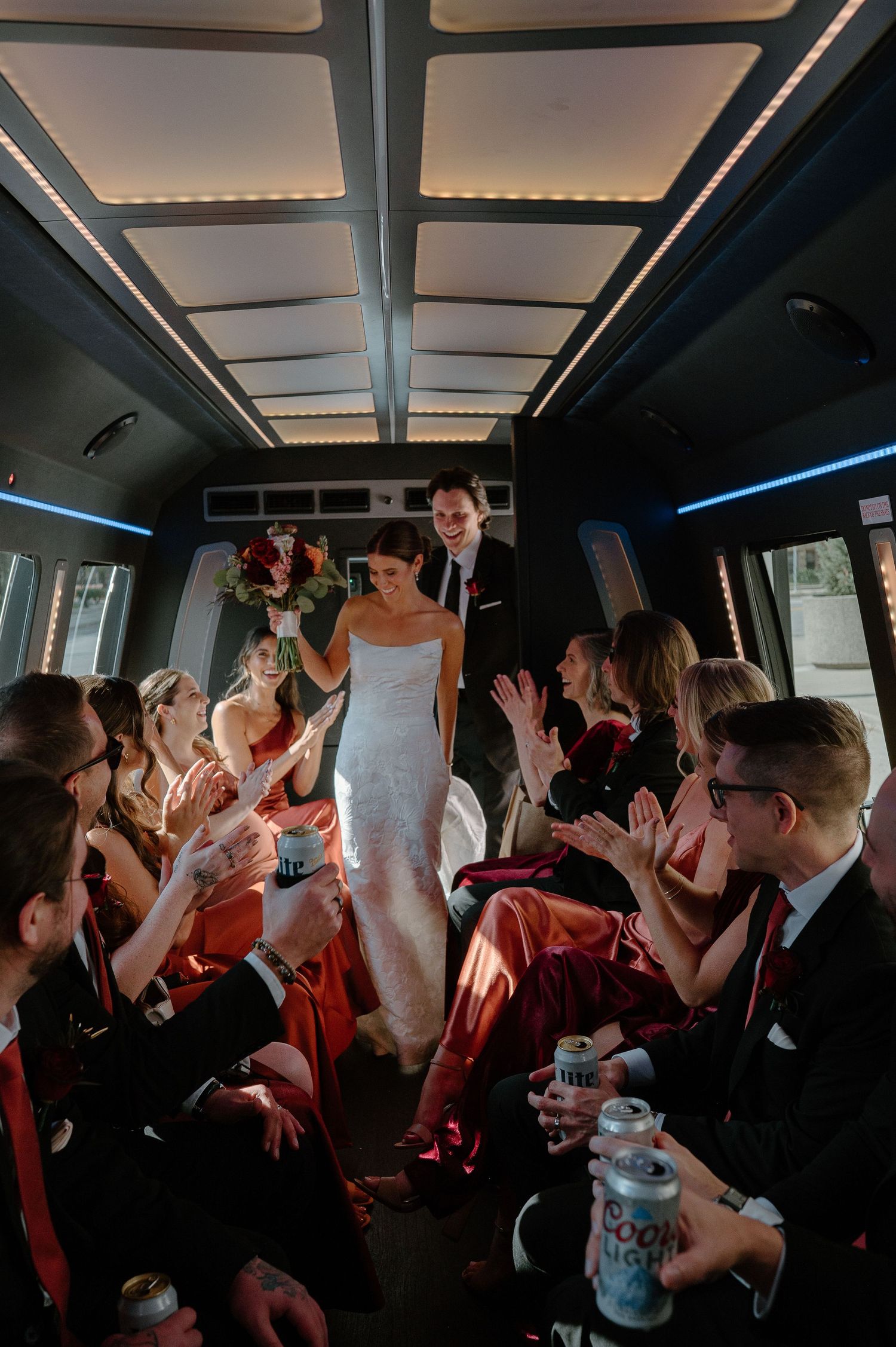 Wedding party celebrates and dances together in a dimly lit party bus with their hands raised in excitement.