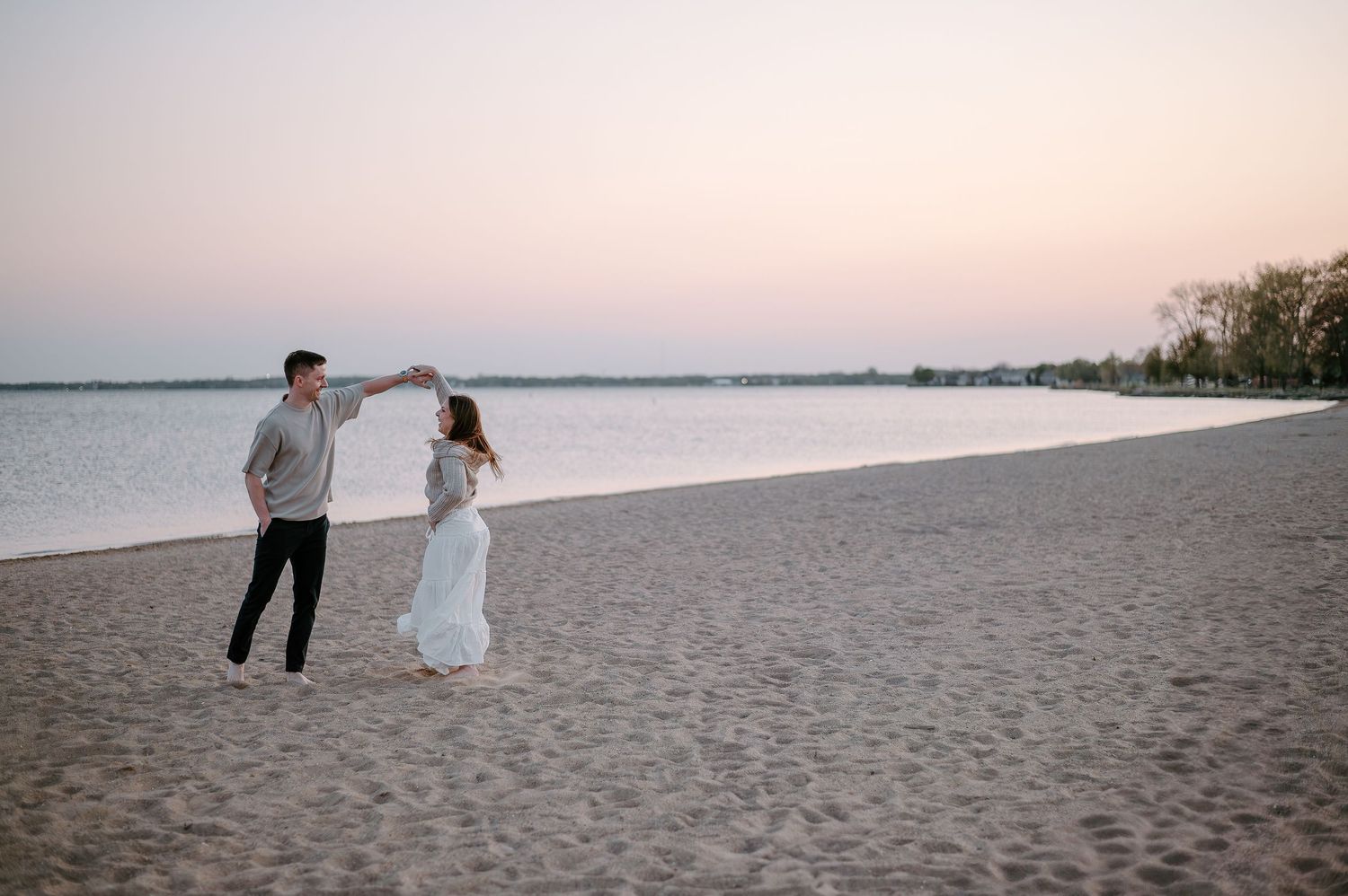 A couple walking along a sandy beach at sunset with a pastel sky.