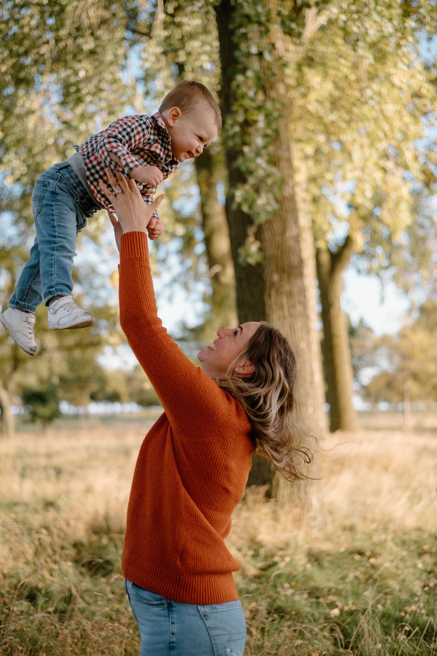 Parent lifting and playing with small child in orange sweater outdoors.