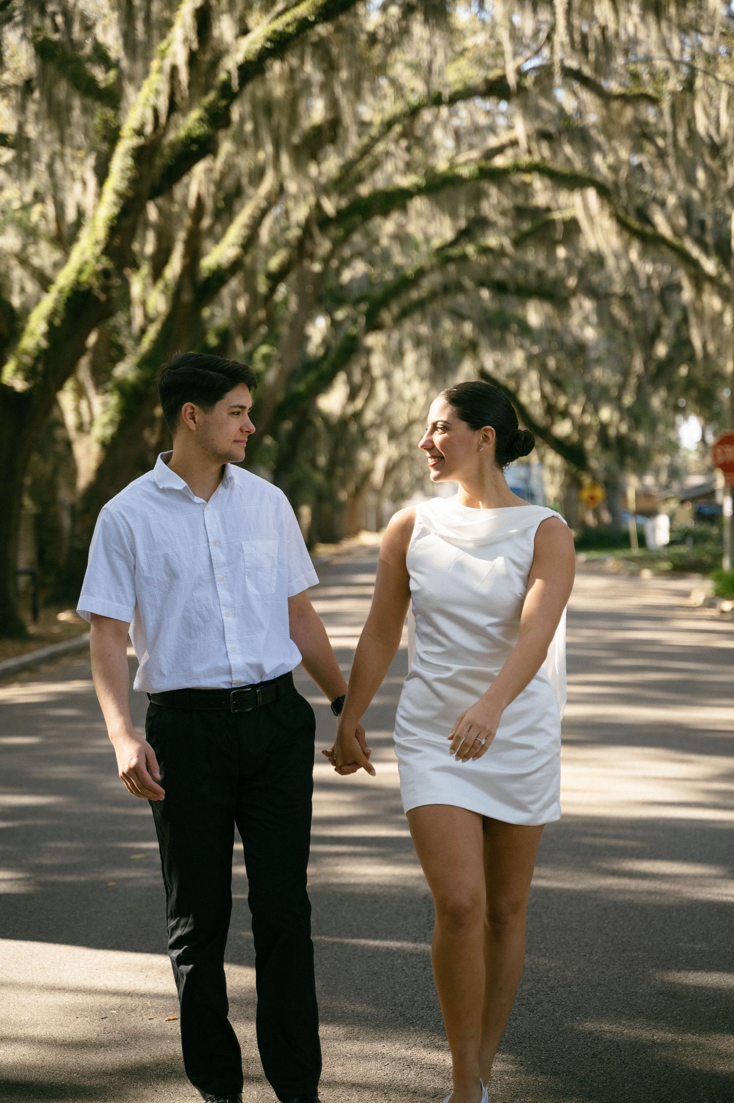 Couple holding hands walking on a tree-lined road, man in white shirt and woman in white mini dress.