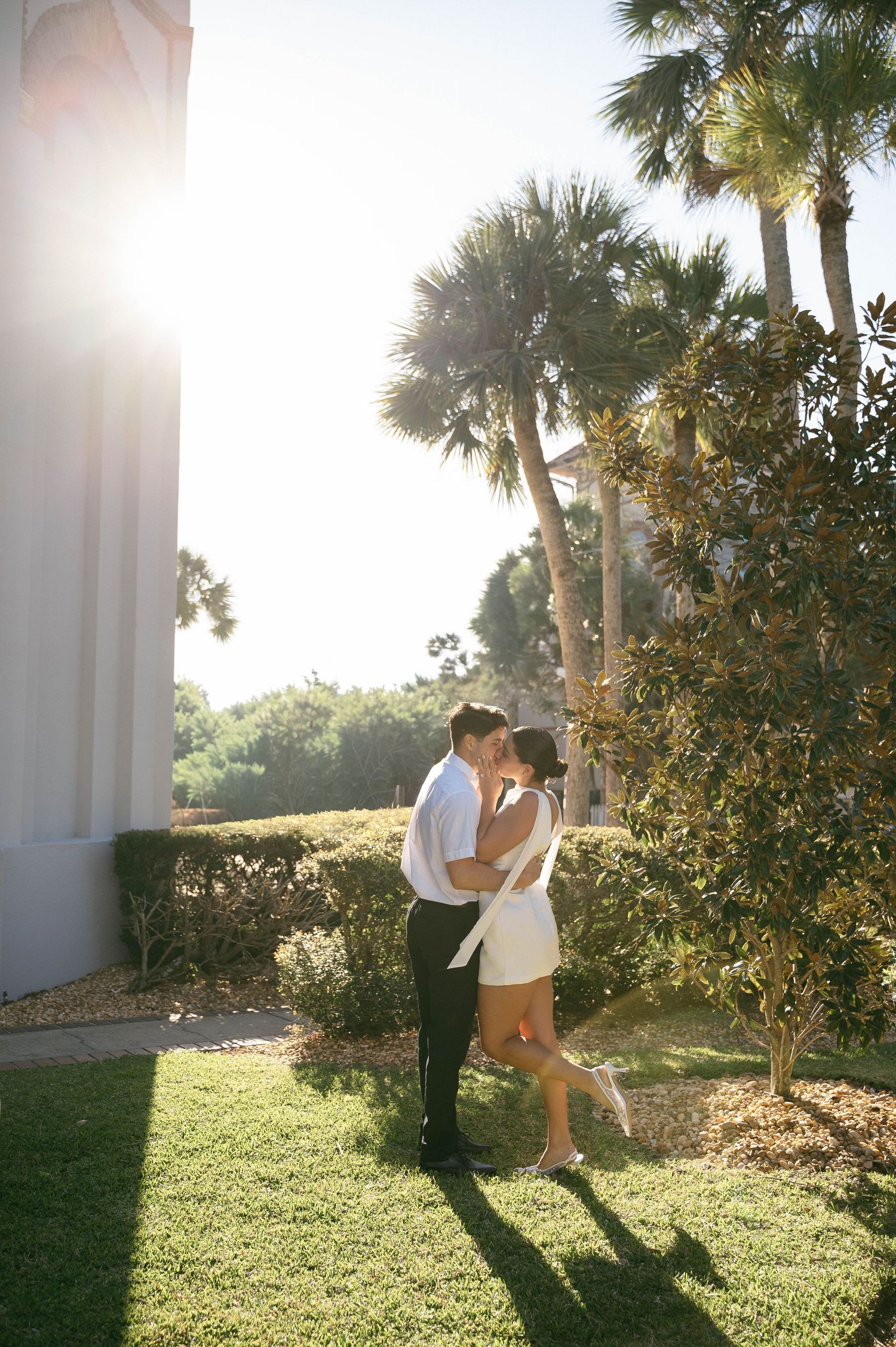 Couple sharing a romantic kiss outdoors on green grass, surrounded by palm trees and lush greenery in warm sunlight.