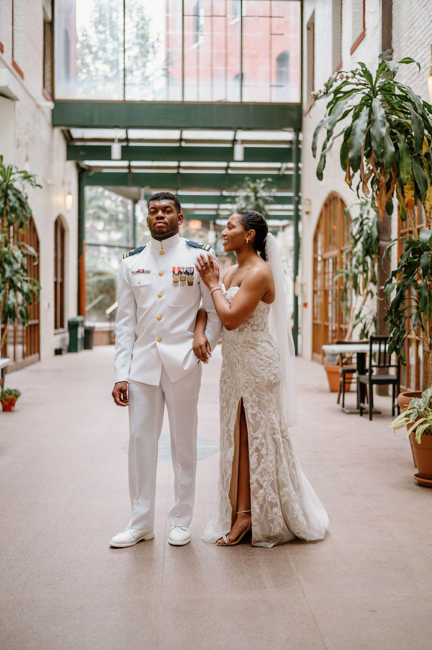 A couple in wedding attire playfully interacts in an indoor corridor with brick columns.