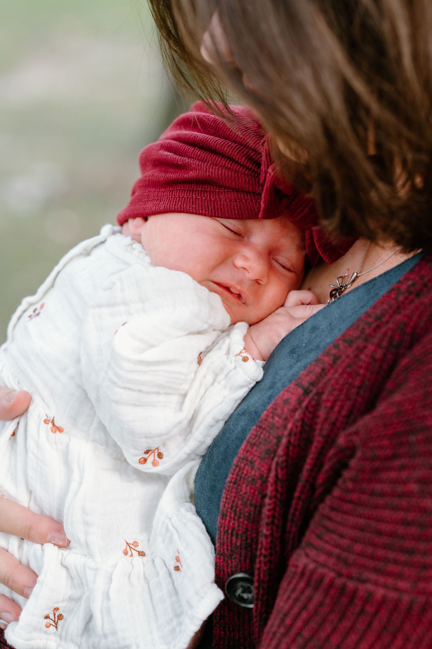 A small infant in a red knit cap and white dress sleeps while being cradled.