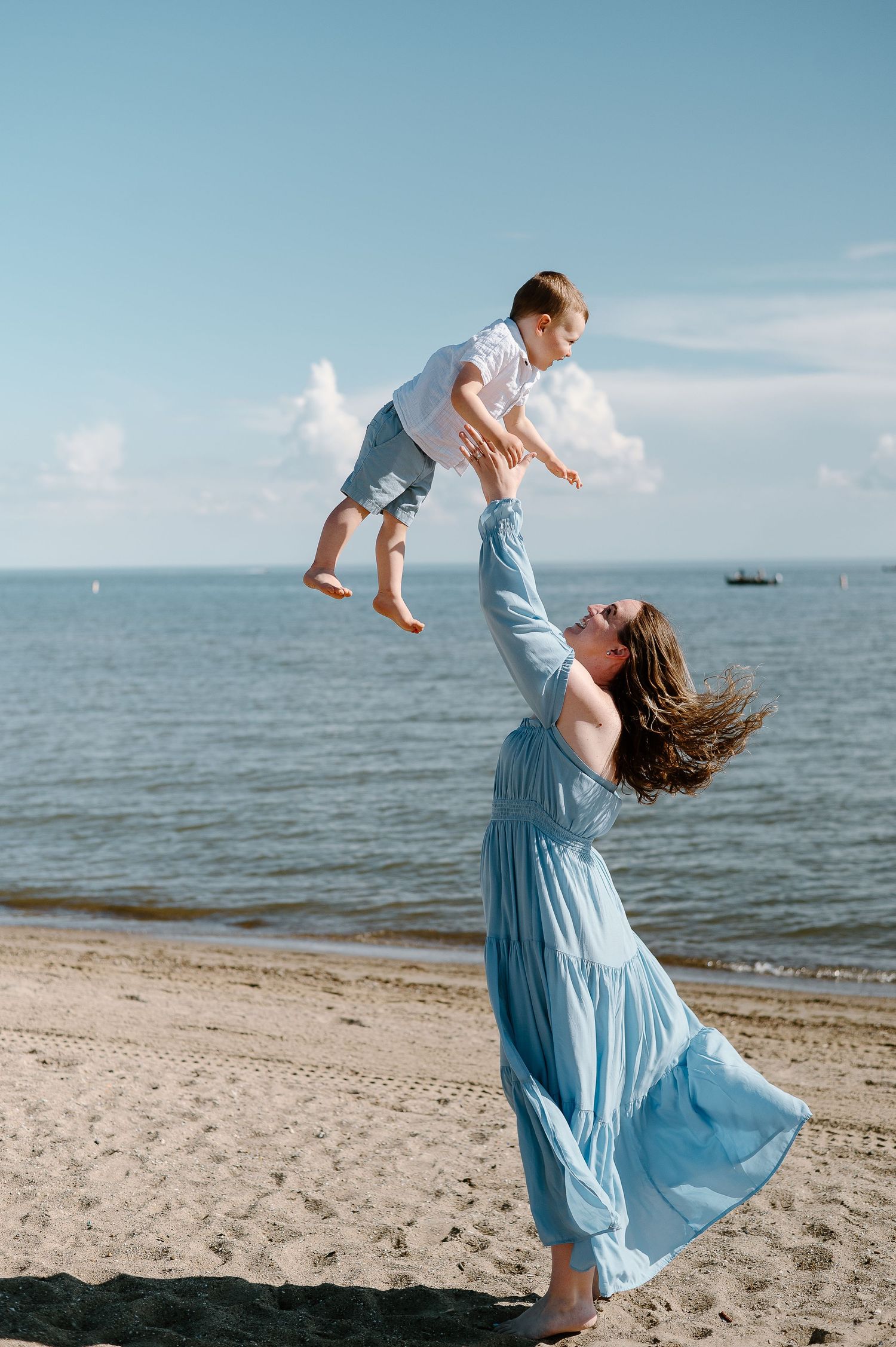 Mother in blue dress lifting child playfully on beach with ocean in background.