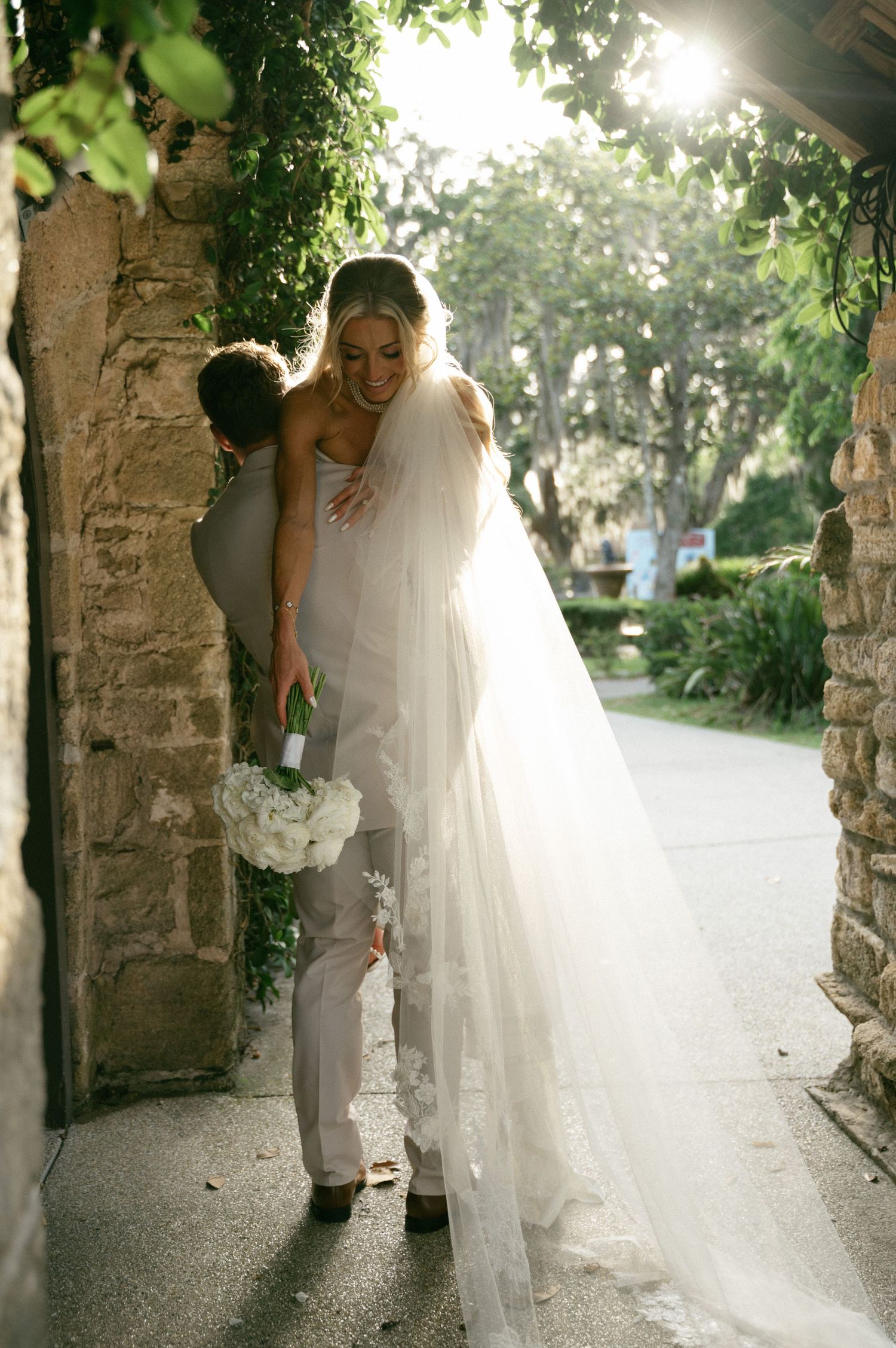 Bride in white tulle gown and veil embraced by groom in gray suit near stone archway with lush greenery.
